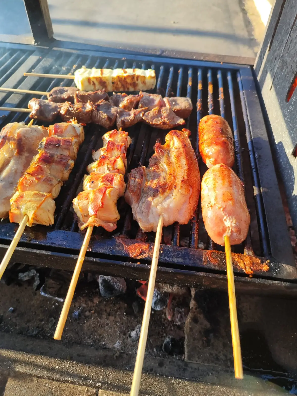 Várias carnes e salsichas assando em churrasco com espetos em grelha ao ar livre.