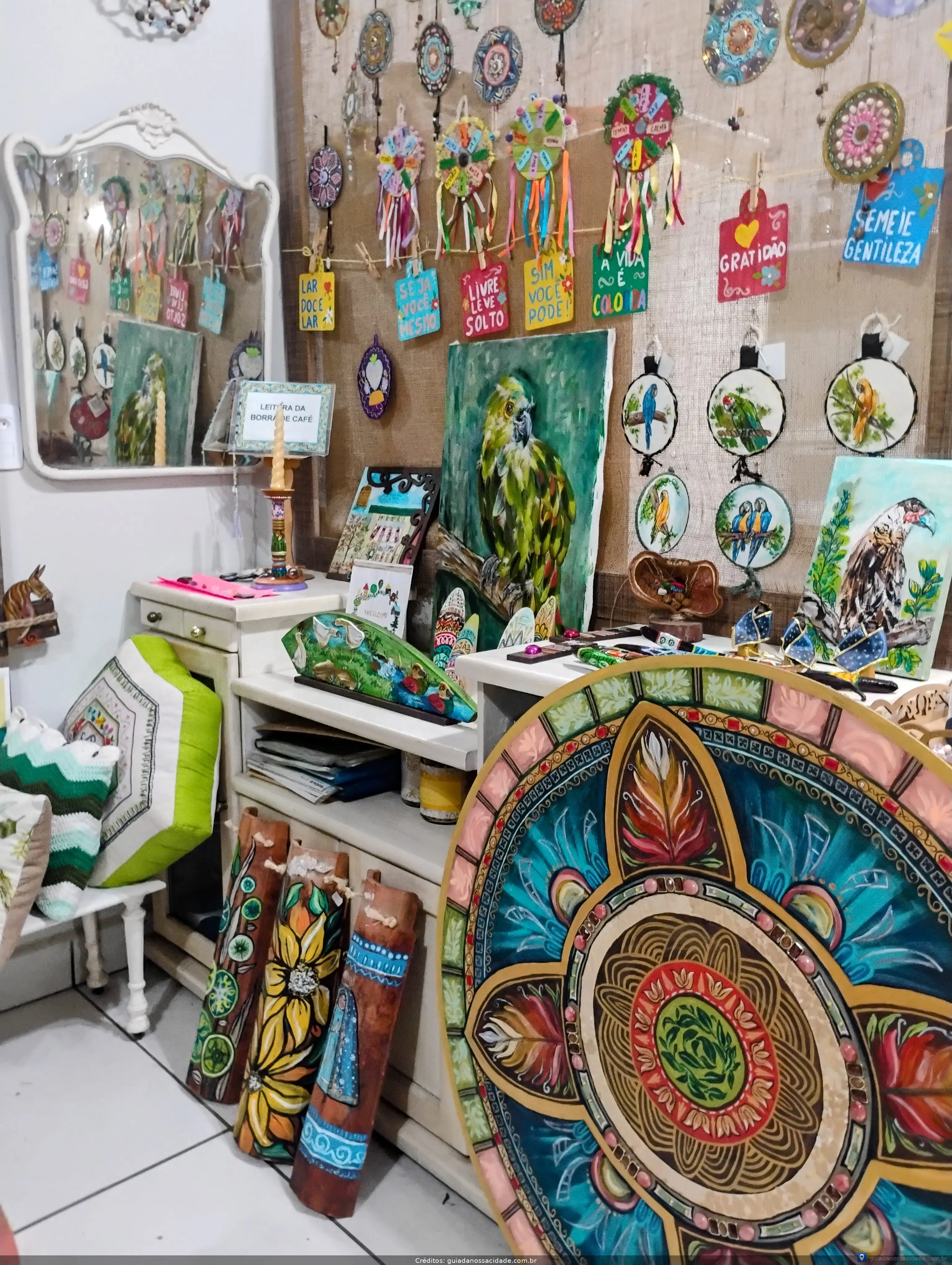 Loja de artesanato com pinturas de arara, mandalas, almofadas coloridas e outros objetos decorativos.