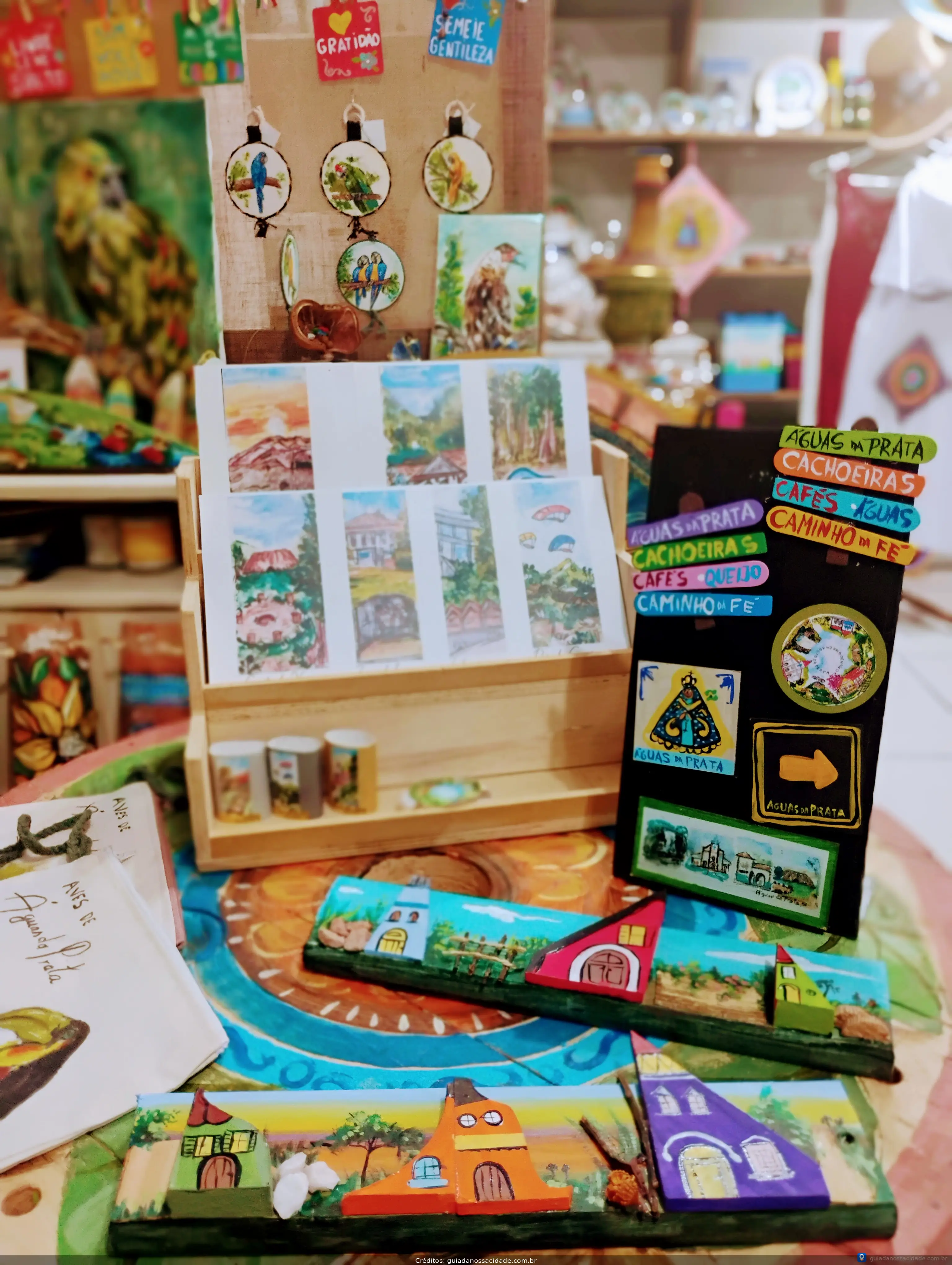 Produtos artesanais coloridos, incluindo pinturas, lembranças e ímãs, exibidos sobre uma mesa em uma loja.