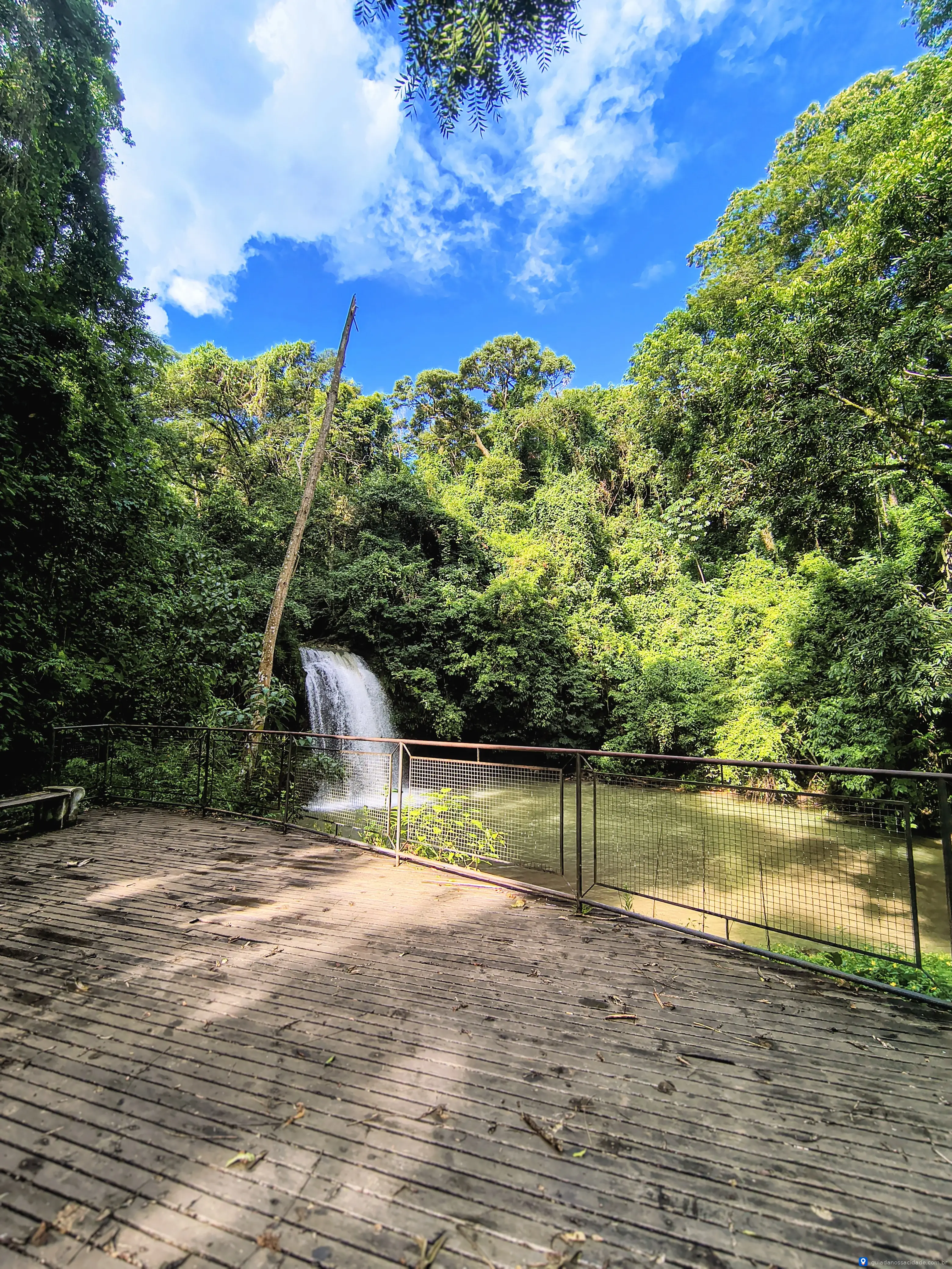 Cachoeira cercada por vegetação densa com plataforma de madeira em dia ensolarado.