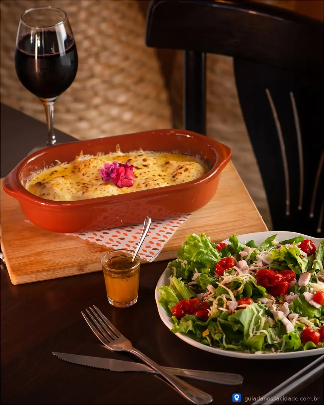 Prato de lasanha gratinada ao lado de salada verde com tomate cereja e taça de vinho tinto.
