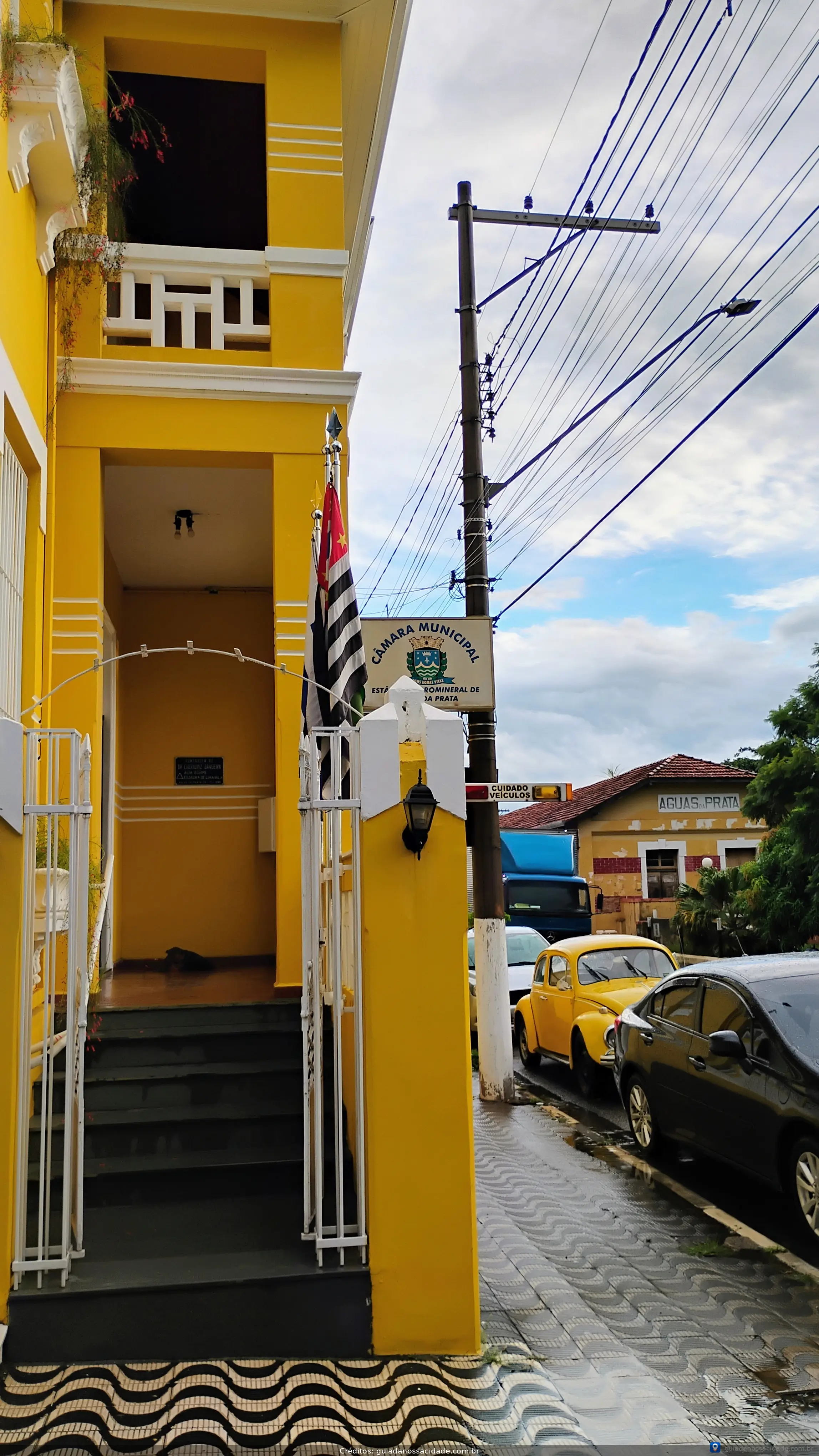Entrada de prédio amarelo com bandeira de São Paulo, placa de Câmara Municipal e carros estacionados na calçada.