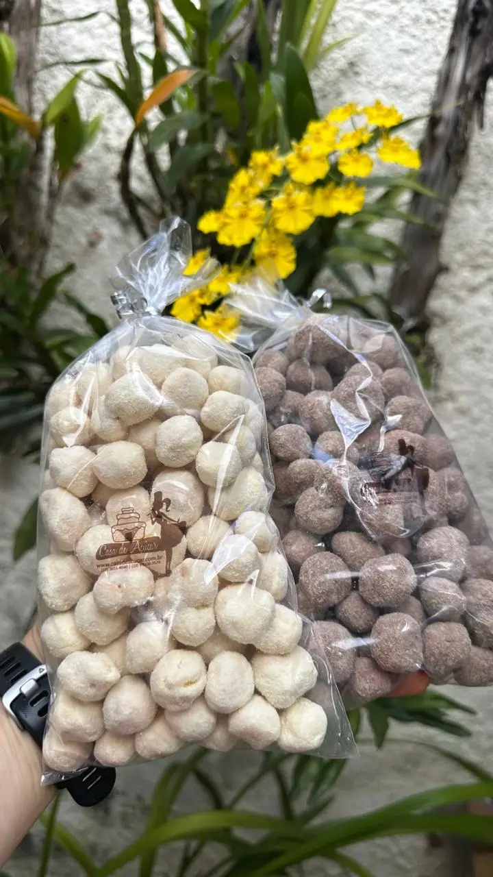 Duas sacolas de biscoitos crocantes, uma branca e outra marrom, seguras por uma mão, com flores amarelas ao fundo.