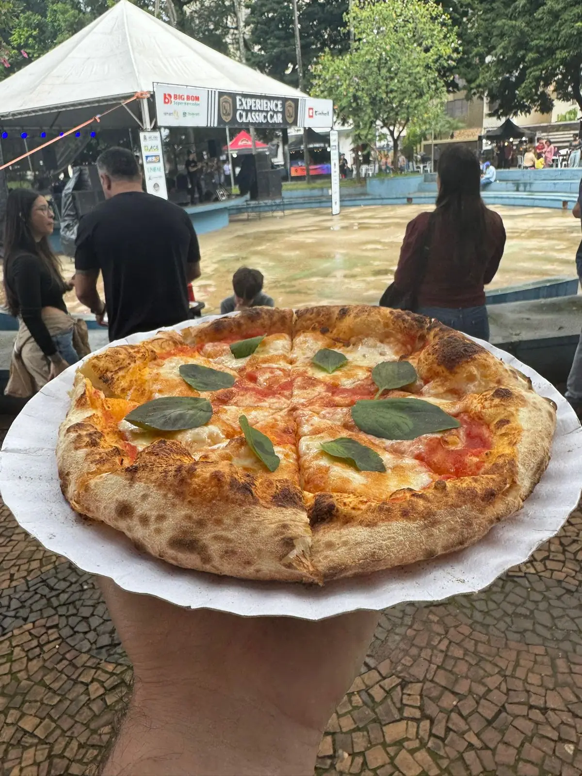 Pizza margherita em mãos na frente de palco em evento ao ar livre.