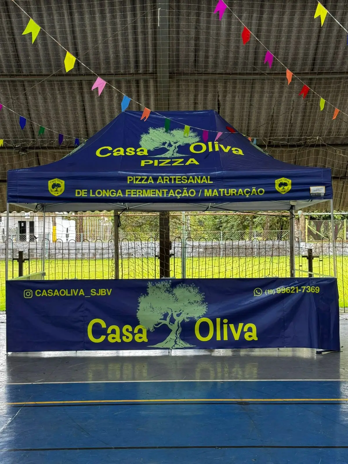 Tenda azul com logotipo Casa Oliva e faixa de pizza artesanal em quadra esportiva coberta.