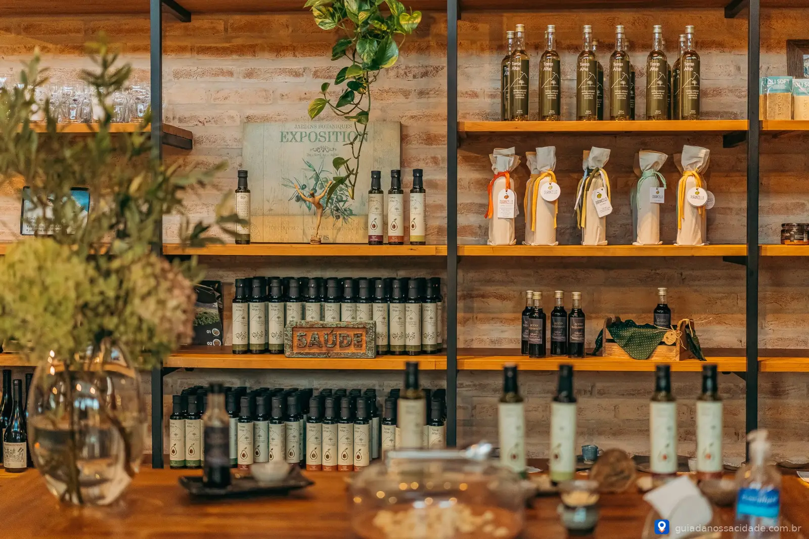 Prateleiras com garrafas de azeite, vinhos embrulhados e decoração rústica em ambiente interno.