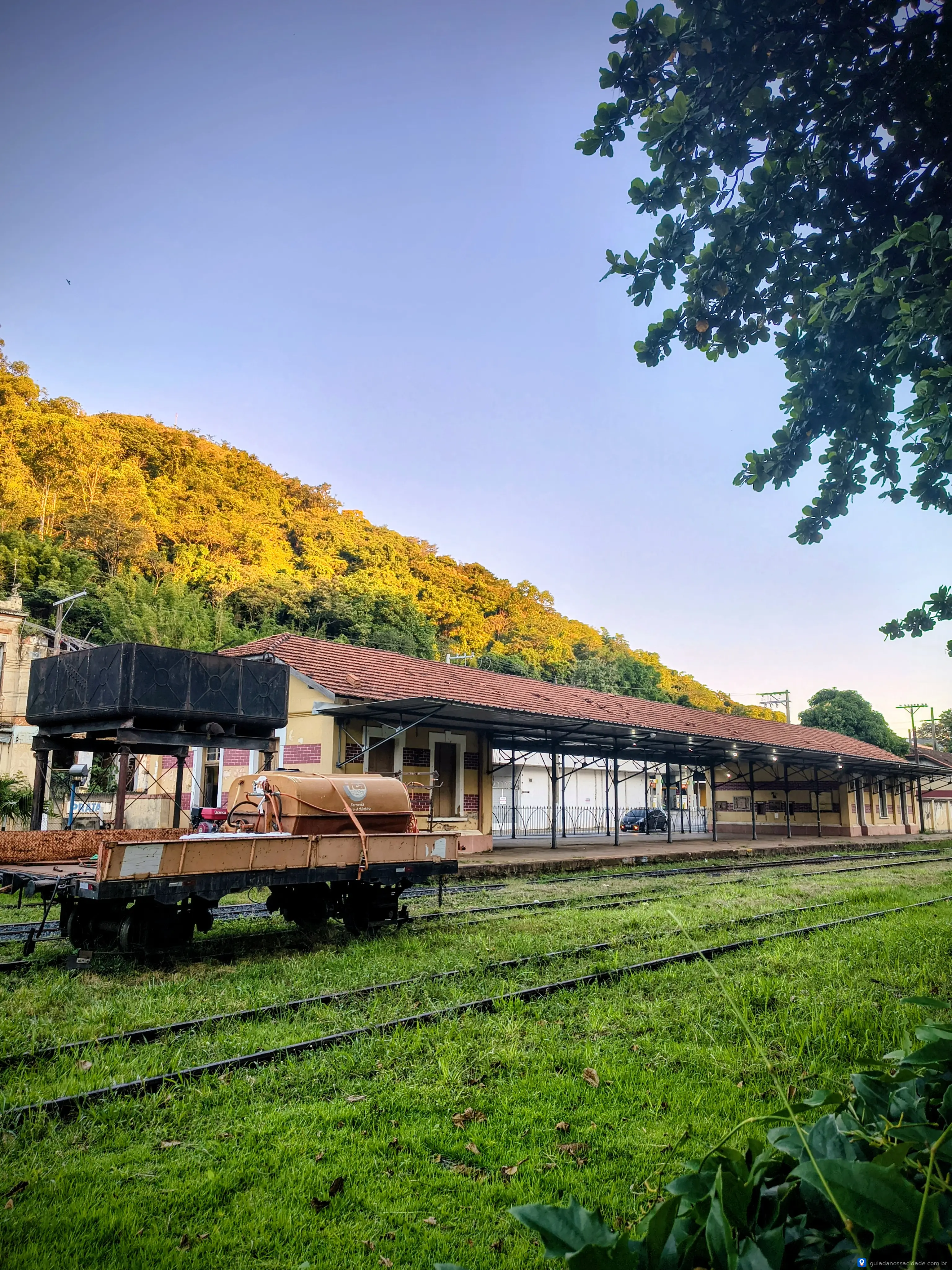 Estação ferroviária antiga com vagão e vegetação ao redor sob céu azul claro.
