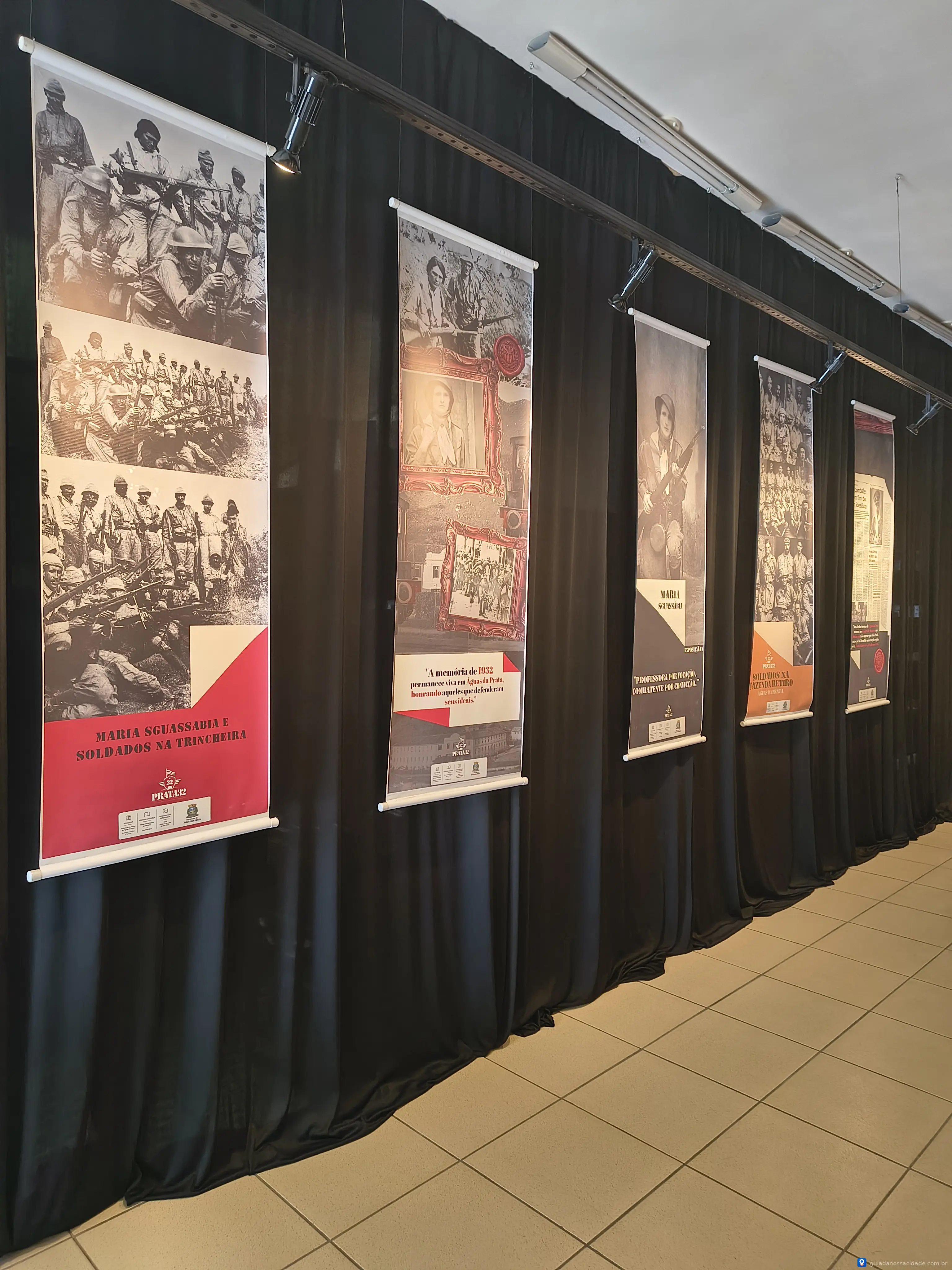 Exposição com seis cartazes de fotos históricas pendurados em uma parede coberta por cortina preta.