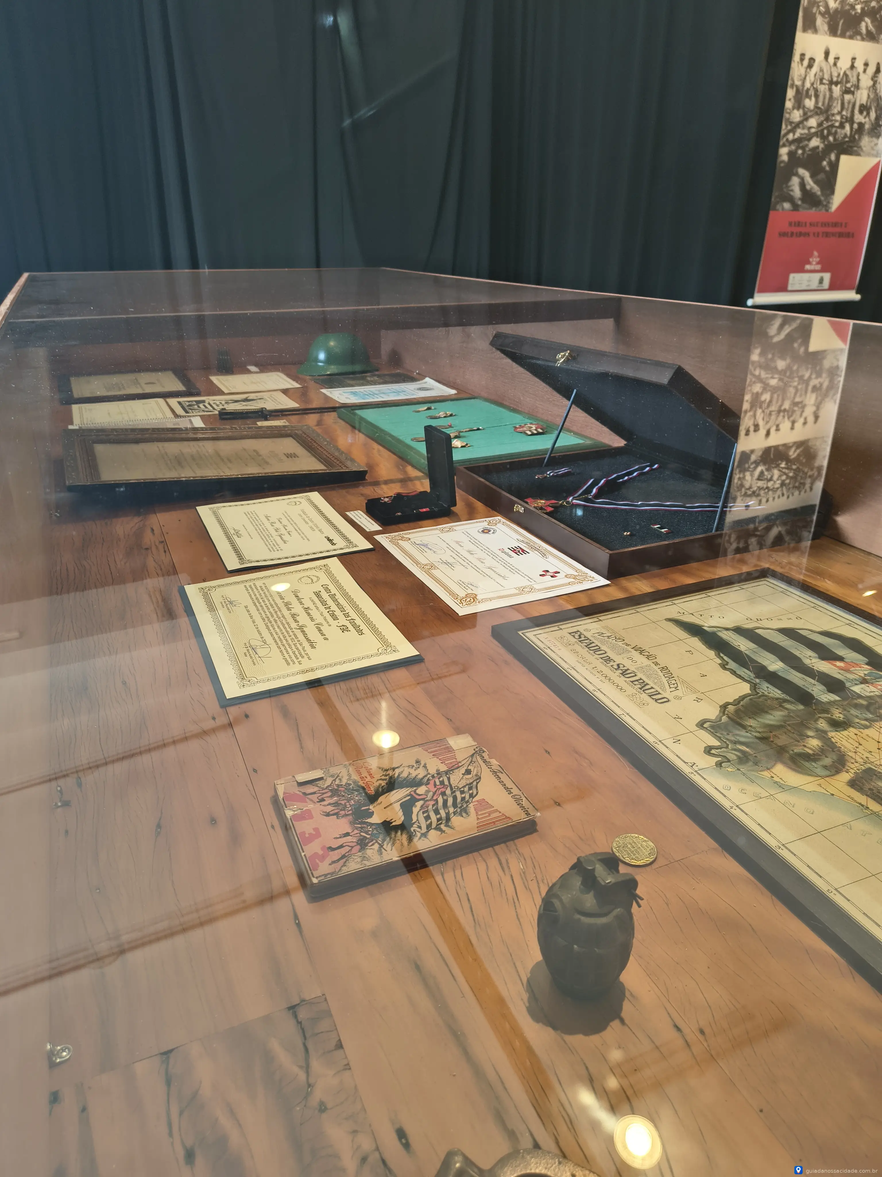 Vitrine com diplomas, mapa, granada decorativa e objetos variados sob vidro em exposição.