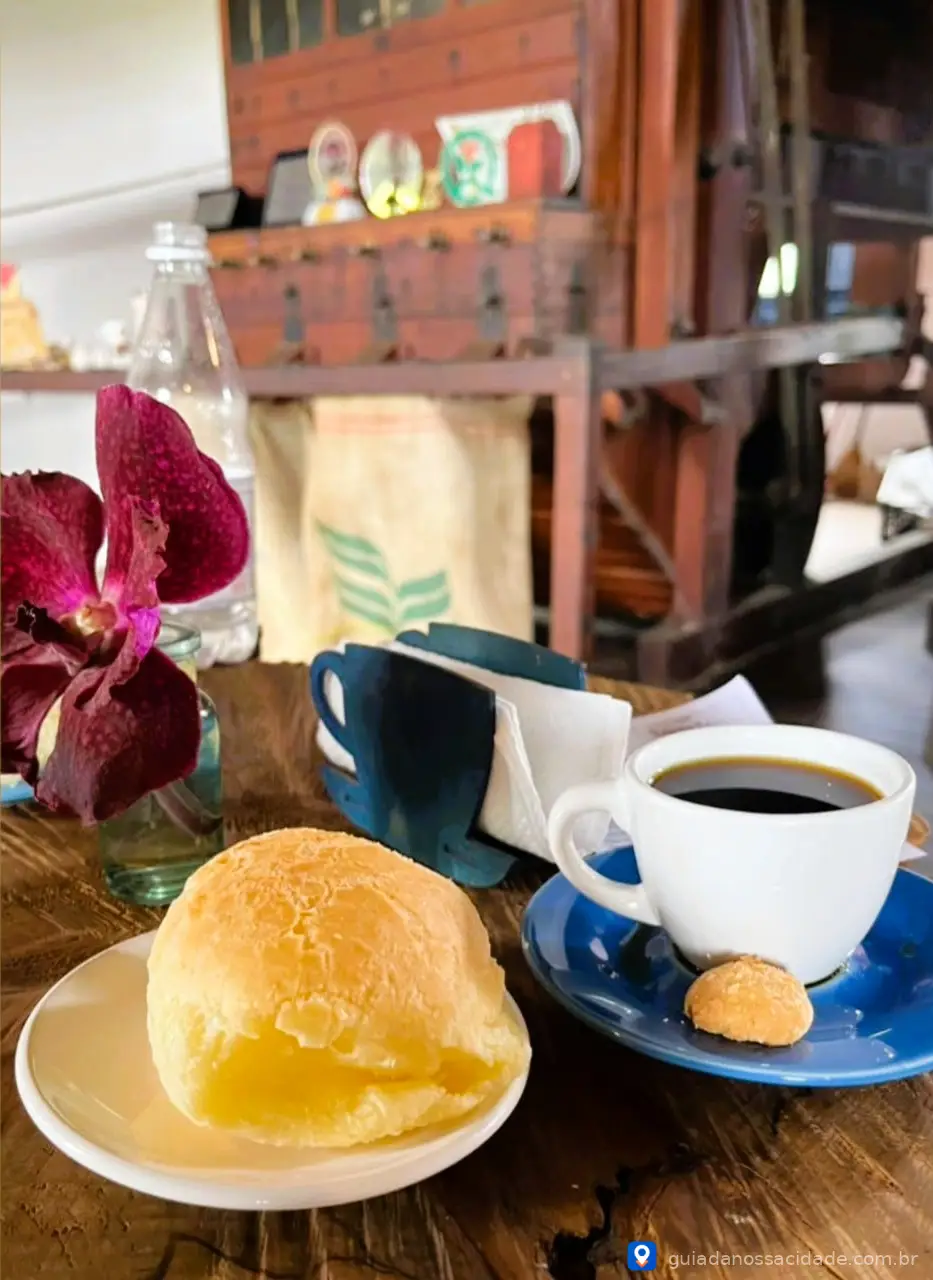 Mesa com xícara de café, pão de queijo em prato, biscoito e garrafa ao lado de uma flor roxa.