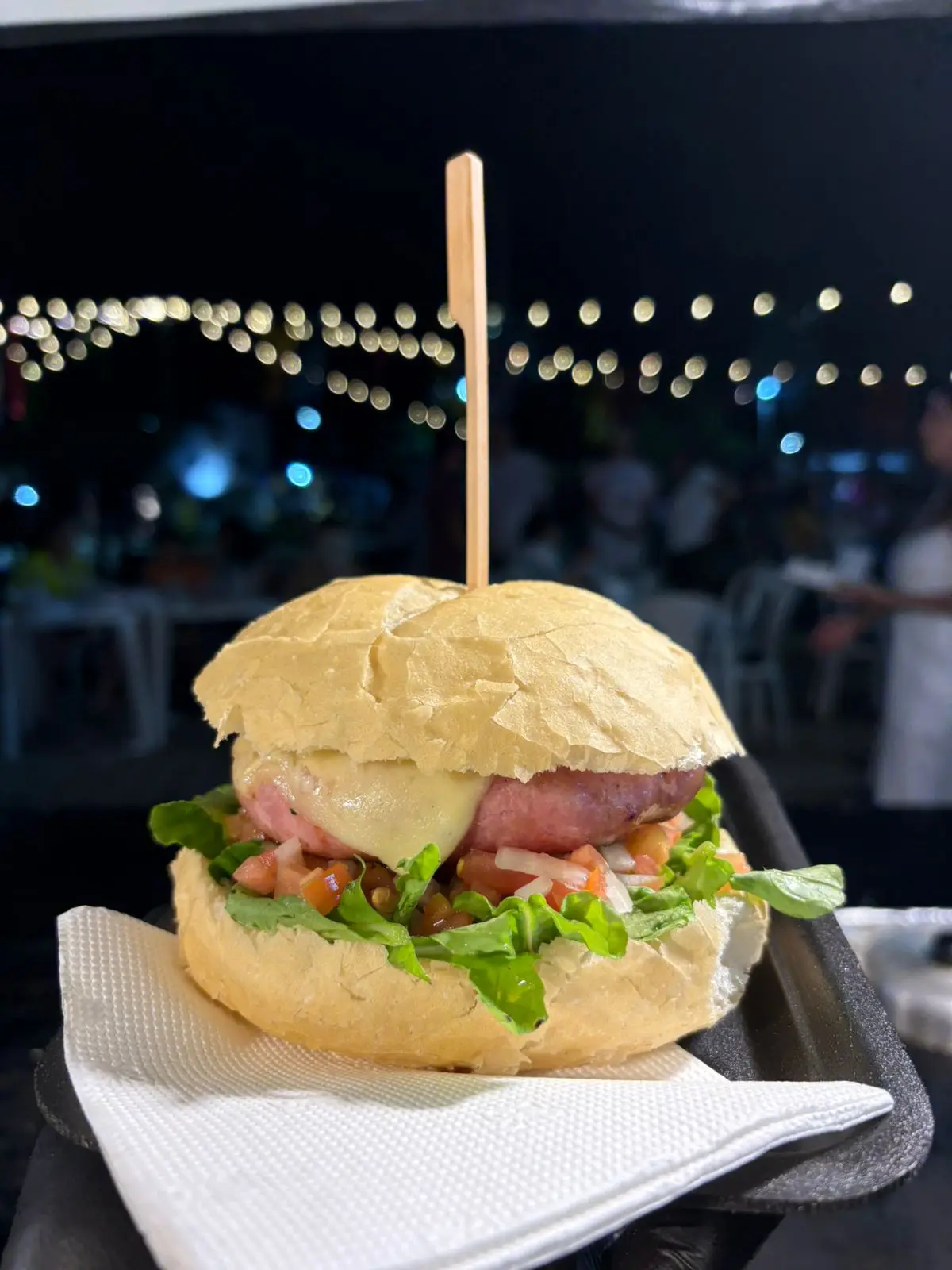Pão de hambúrguer recheado com alface, tomate, queijo derretido e salsicha, decorado com palito de madeira.