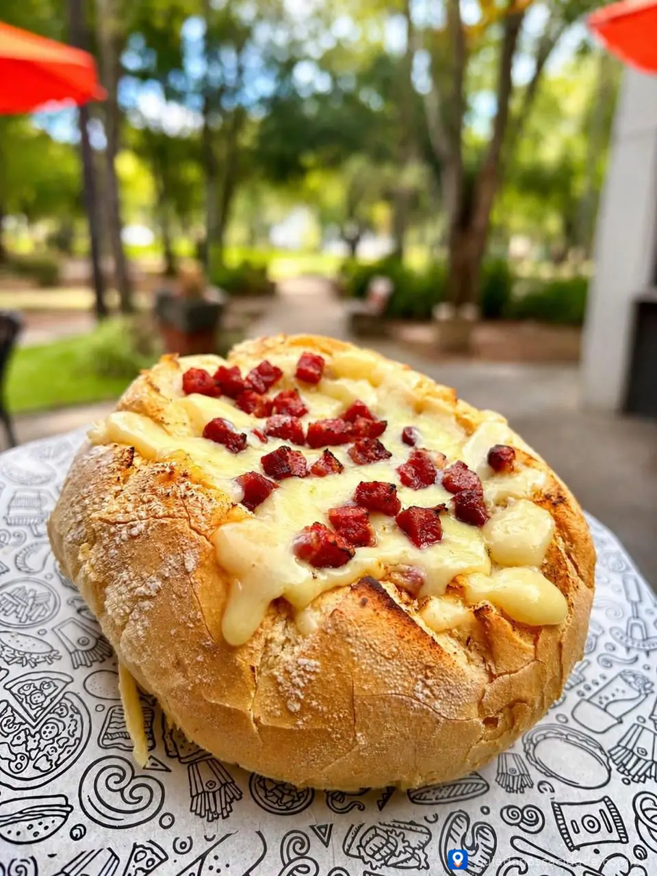 Pão recheado com queijo derretido e pedaços de calabresa sobre papel decorado, fundo desfocado de parque.