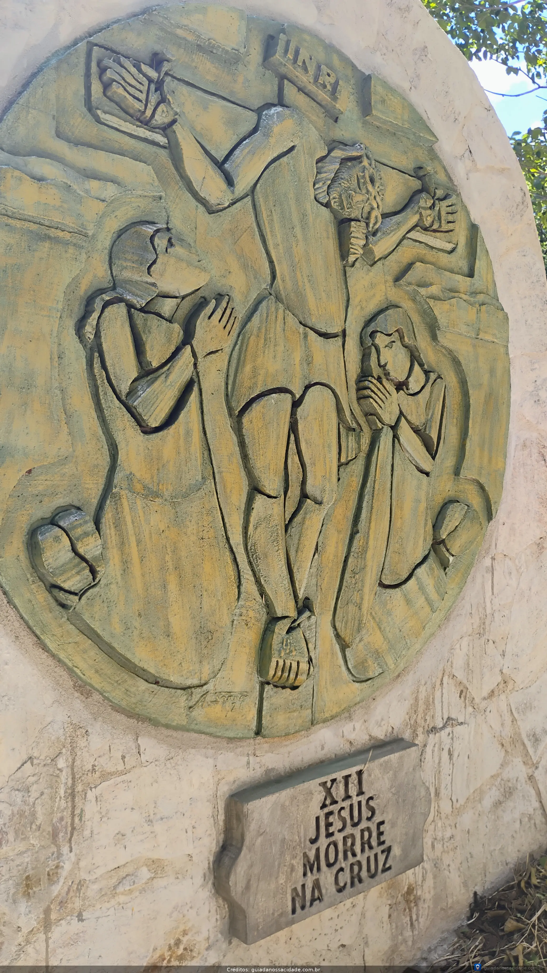 Relevo em pedra de Jesus na cruz com figuras orando ao lado, placa com a inscrição 'XII Jesus morre na cruz'.