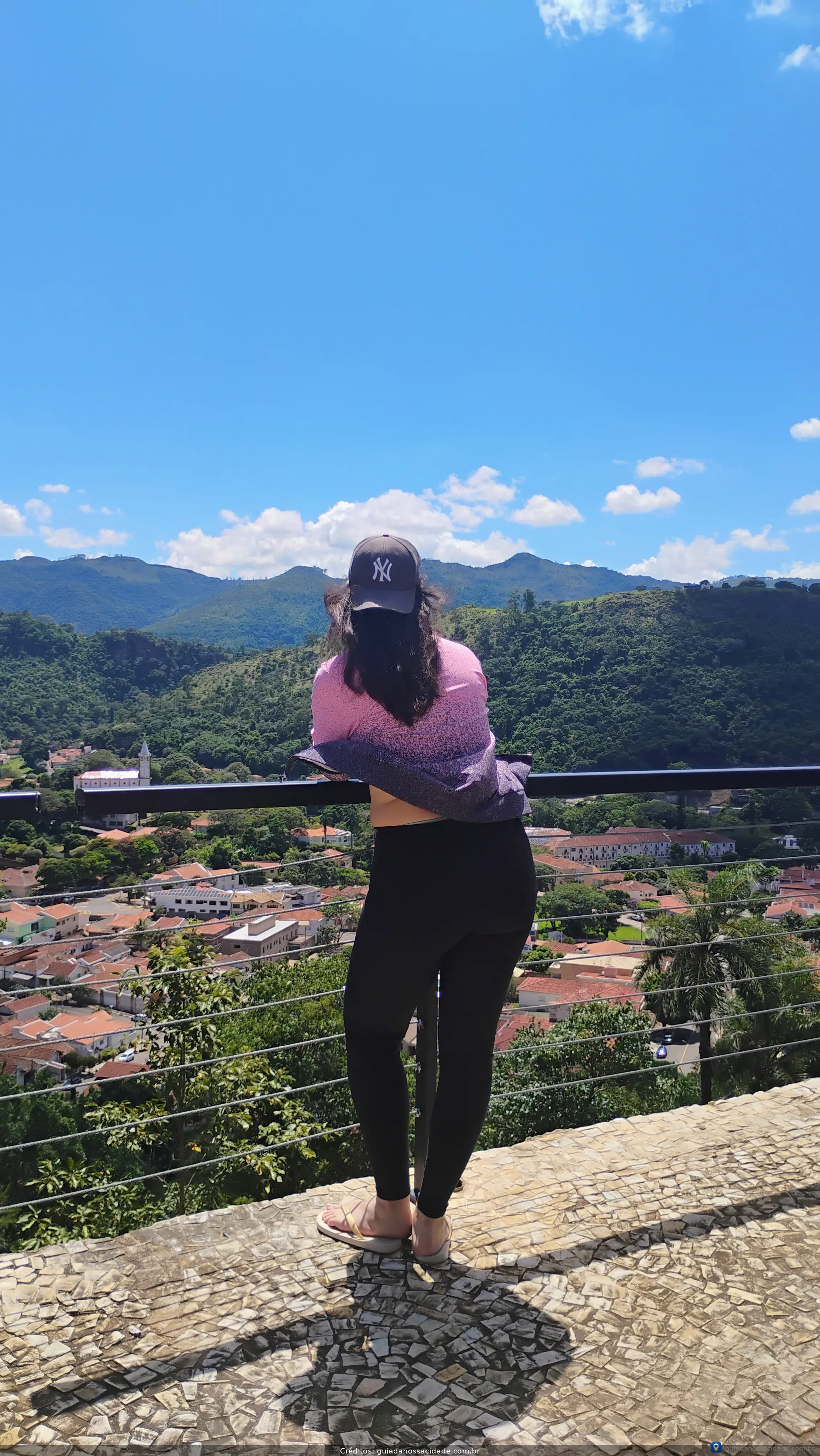 Pessoa de costas com boné observando cidade, cercada por montanhas verdes sob céu azul.