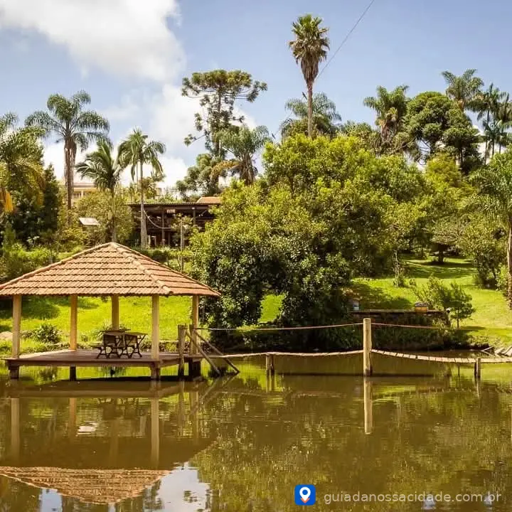 Quiosque de madeira com teto de telhas à beira de um lago rodeado por árvores e palmeiras em um dia ensolarado.