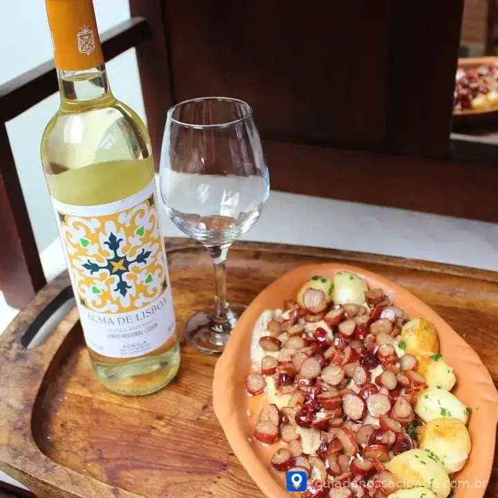 Garrafa de vinho branco, taça vazia, prato de madeira com linguiça em rodelas e batatas.