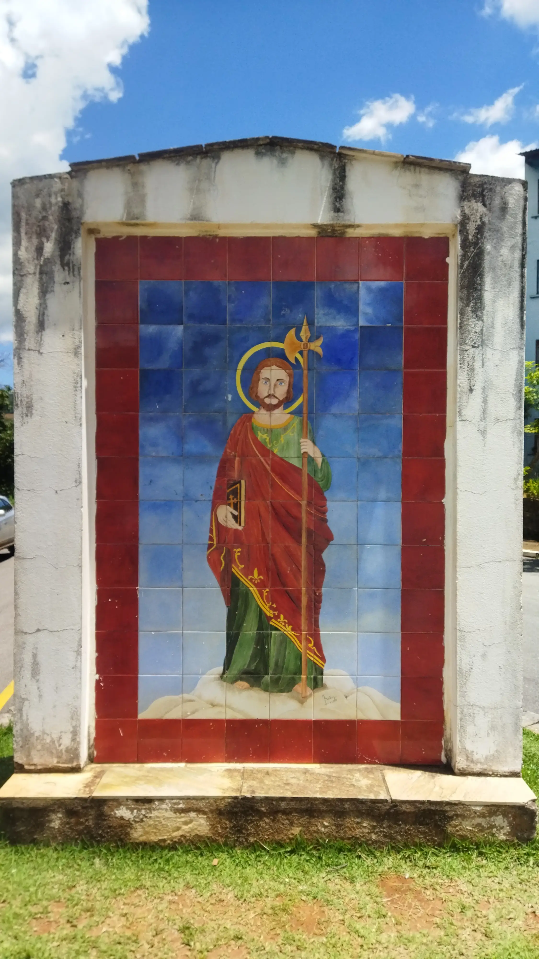Mosaico de santo segurando livro e cetro, vestindo túnica vermelha, em mural externo sob céu azul.