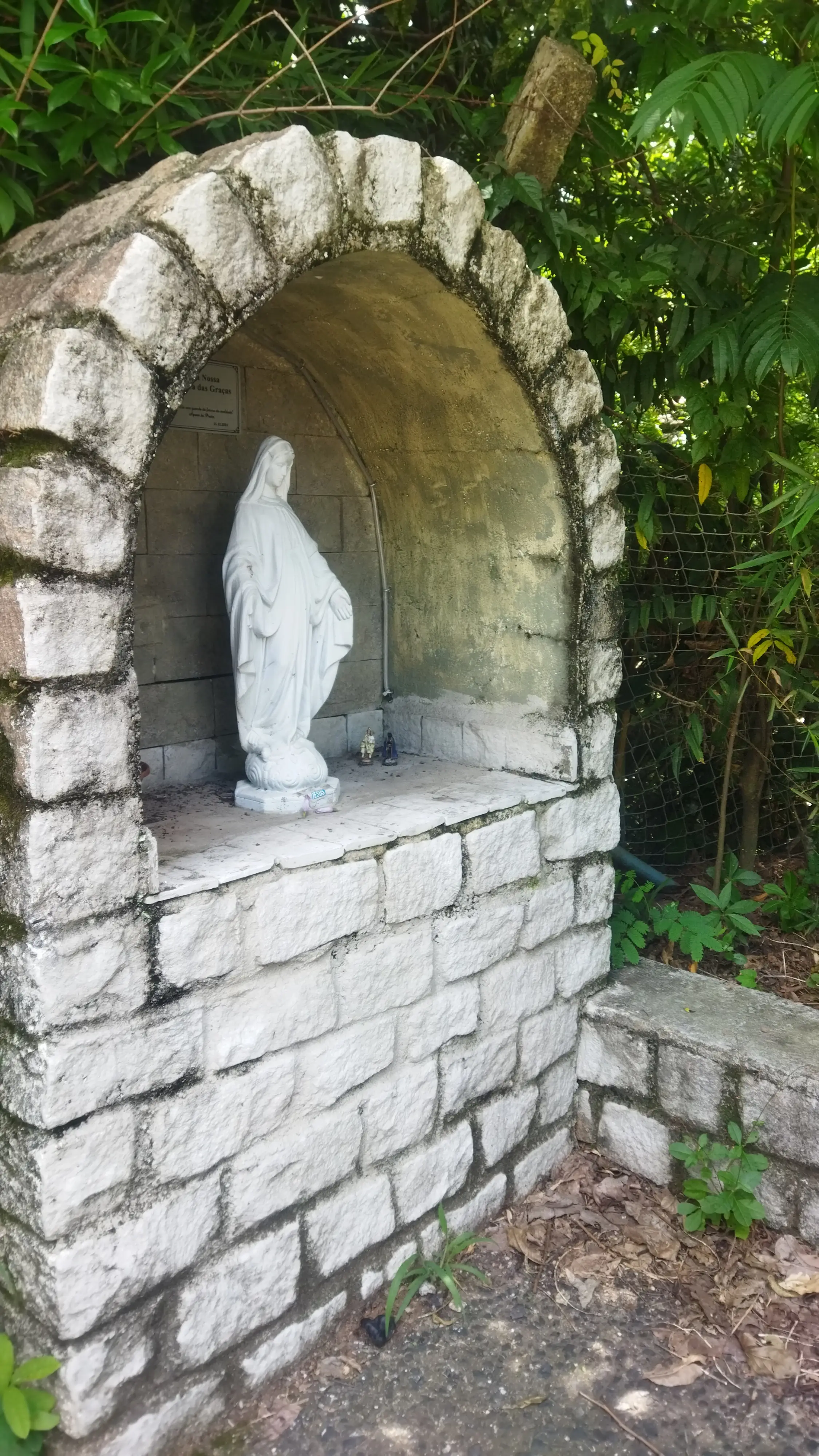 Estátua branca em nicho de pedra com vegetação ao redor.