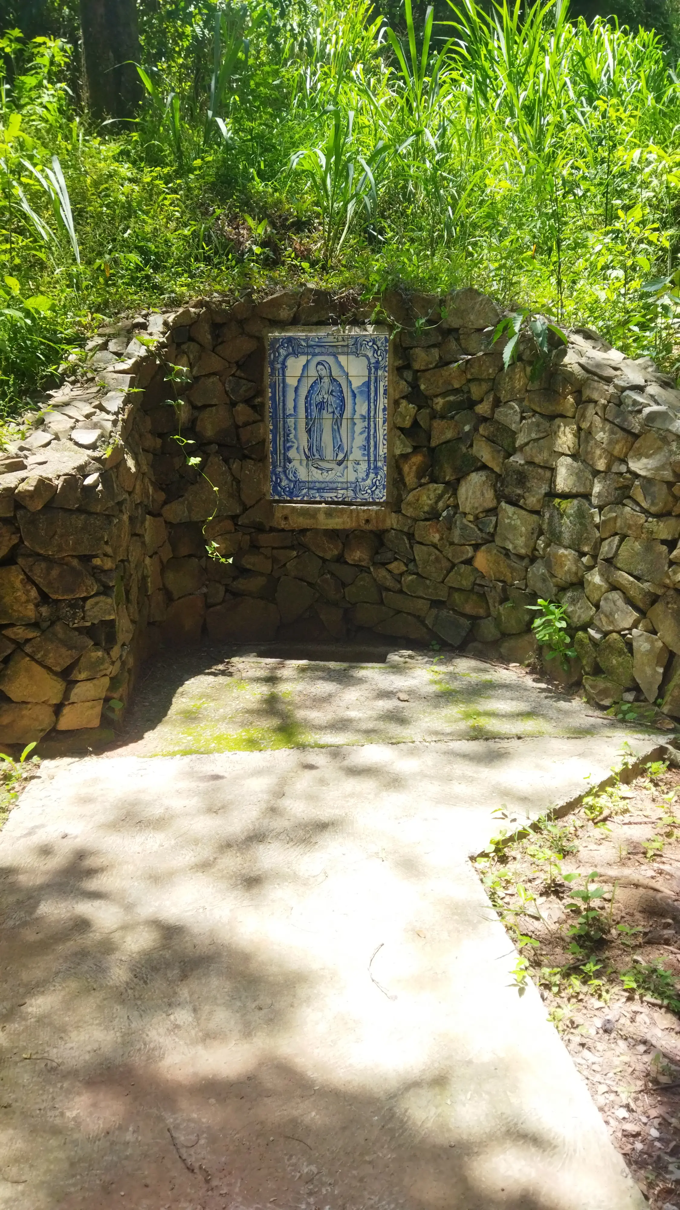 Parede de pedras com azulejo religioso cercada por vegetação verde.
