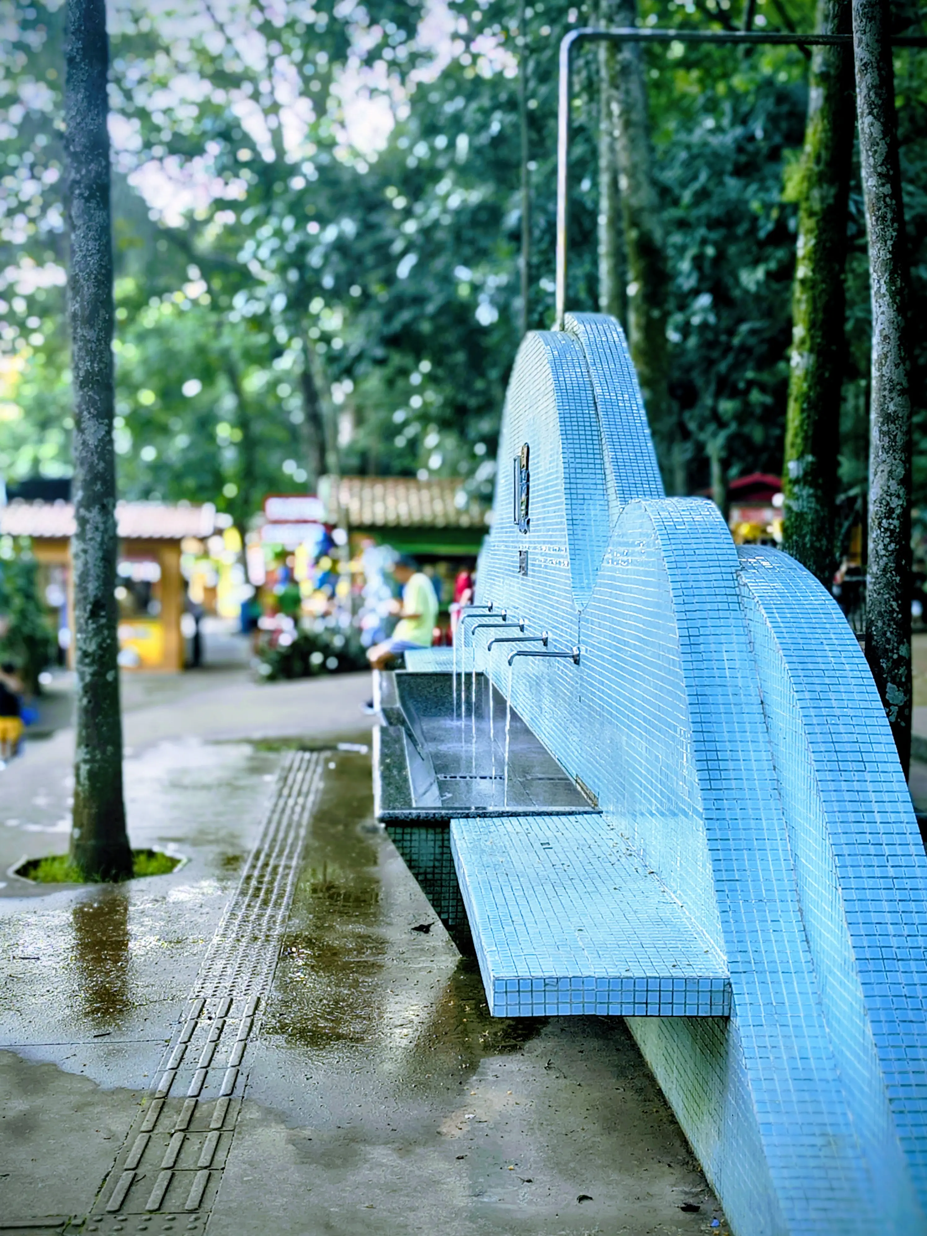 Fonte de água azul em azulejos, com torneiras em um parque arborizado.
