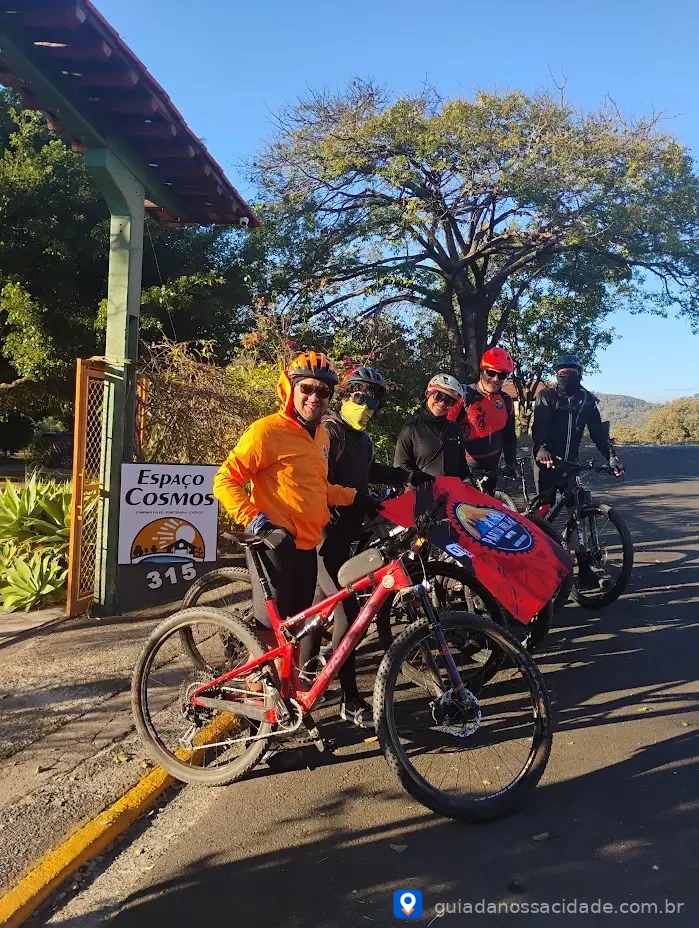 Grupo de ciclistas com capacetes coloridos em frente ao Espaço Cosmos em dia ensolarado.