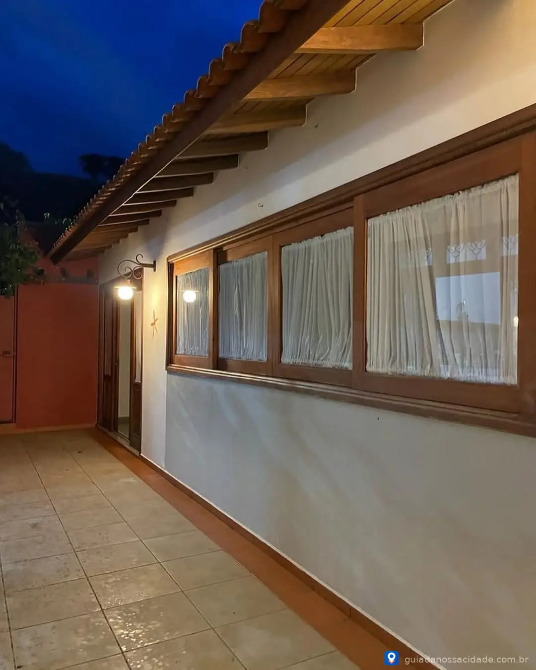 Corredor externo iluminado à noite com coberturas de telhas de madeira e cortinas nas janelas.