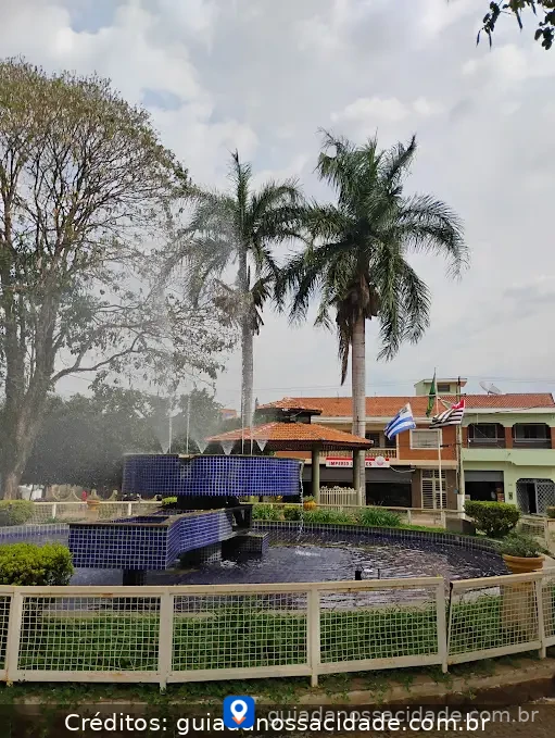 Chafariz azul em praça com palmeiras, bandeiras e edifícios ao fundo.