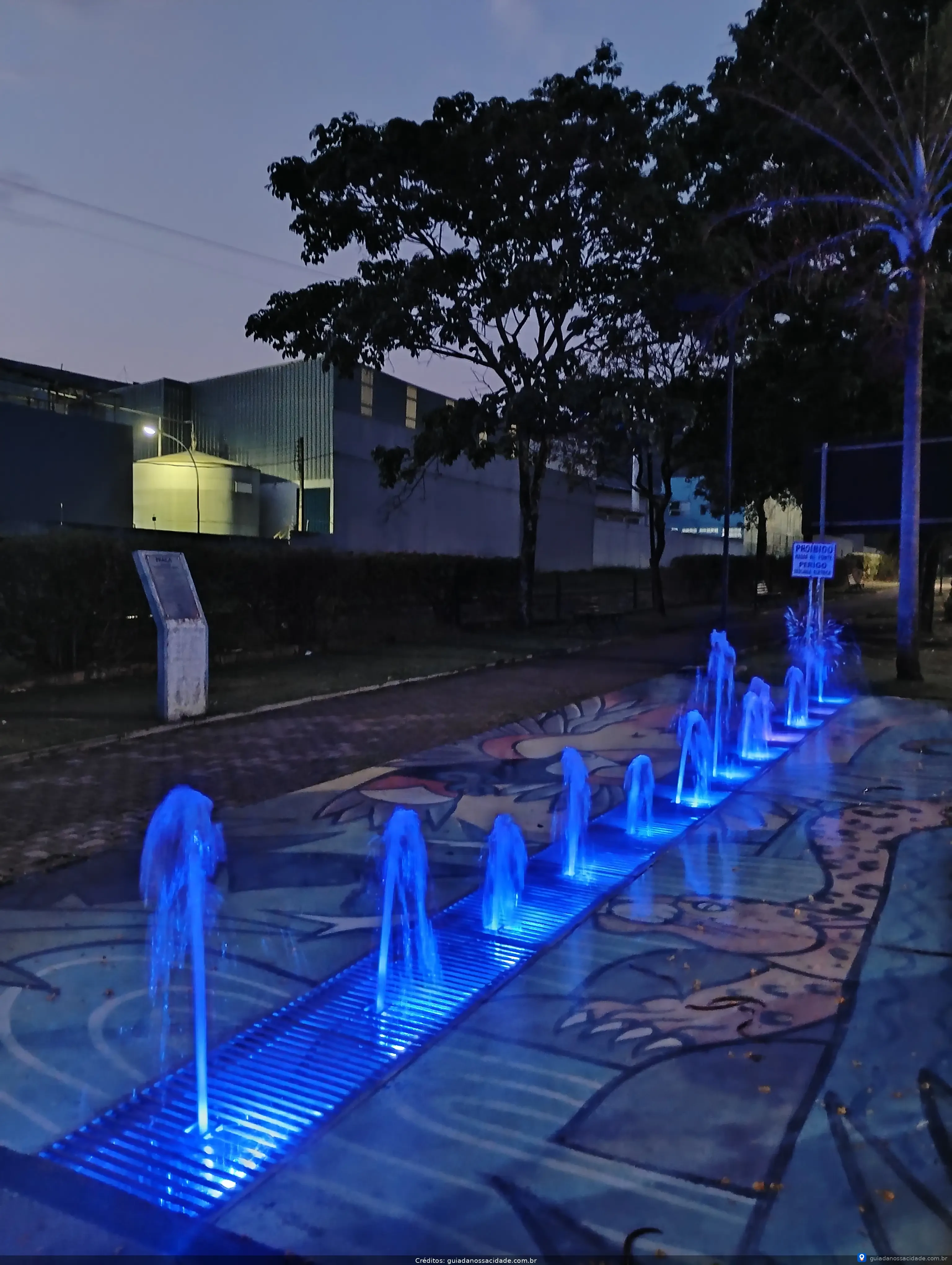 Fontes iluminadas de azul em calçada com árvores ao fundo durante o anoitecer.