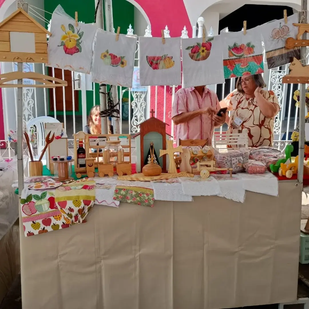 Banca de feira com artesanatos de madeira, toalhas bordadas e pessoas conversando ao fundo.