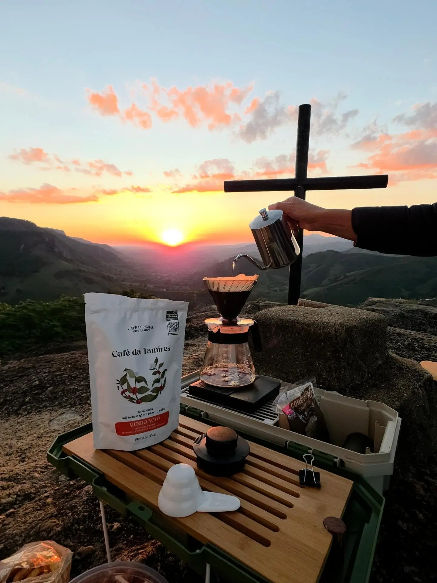 Mesa de café ao pôr do sol com cafeteira, pacote de café e montanhas ao fundo.
