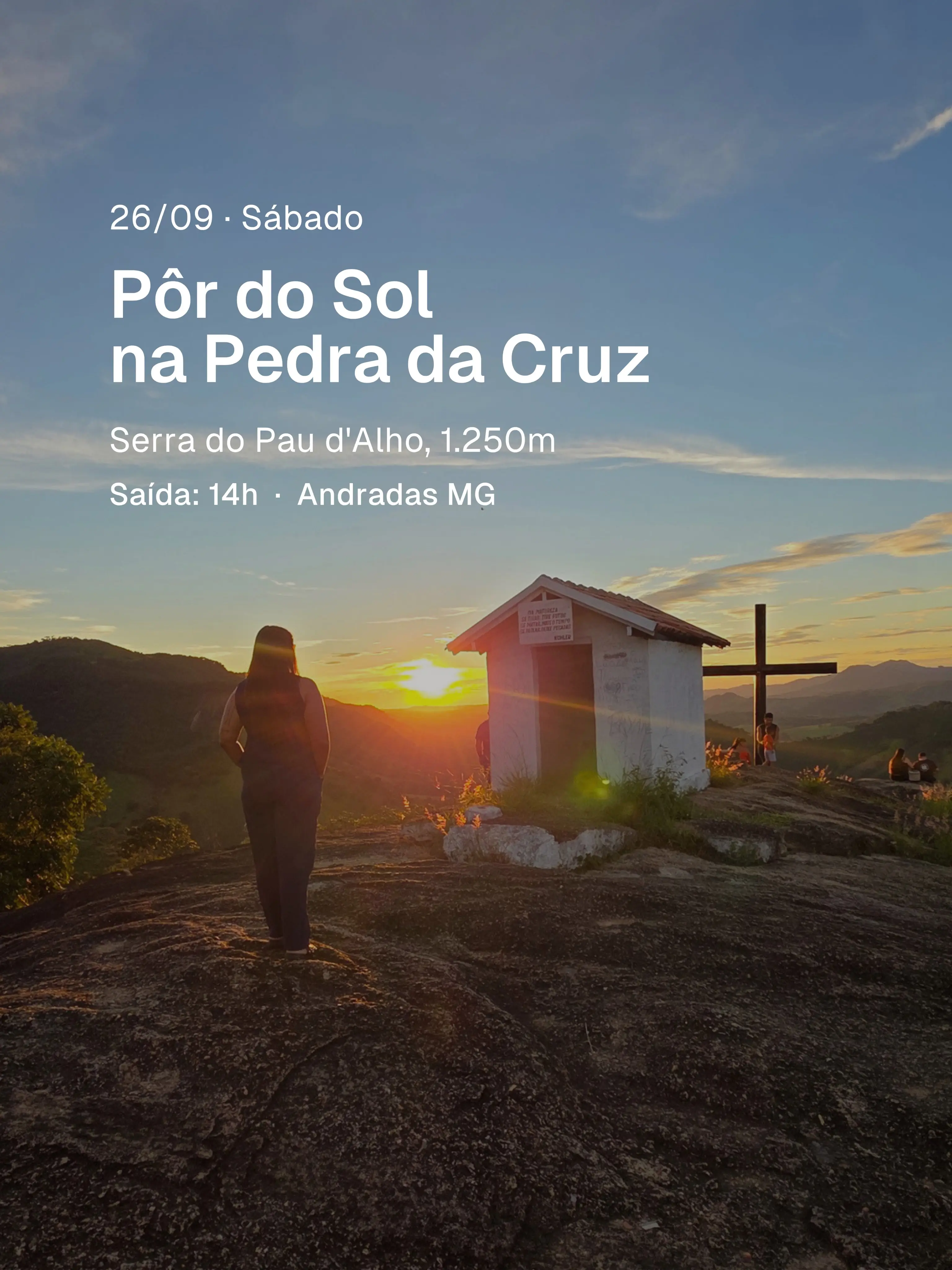 Pessoa observando pôr do sol ao lado de capela e cruz no topo da Pedra da Cruz.