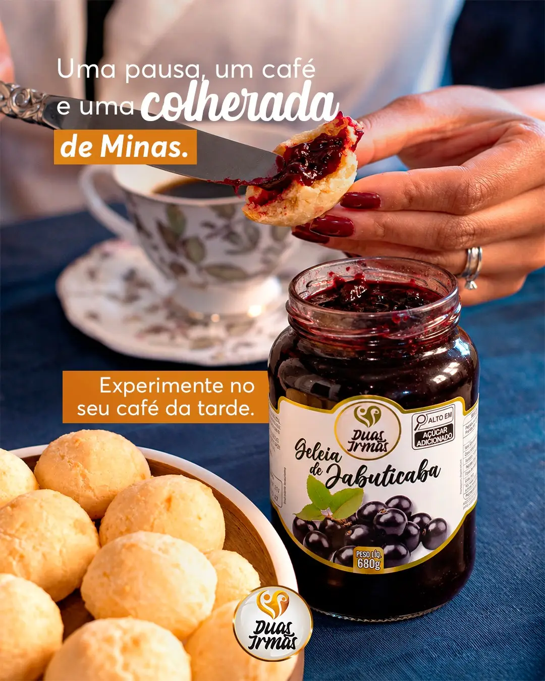 Mão segurando pão de queijo com geleia de jabuticaba, pote de geleia e uma xícara de café ao fundo.