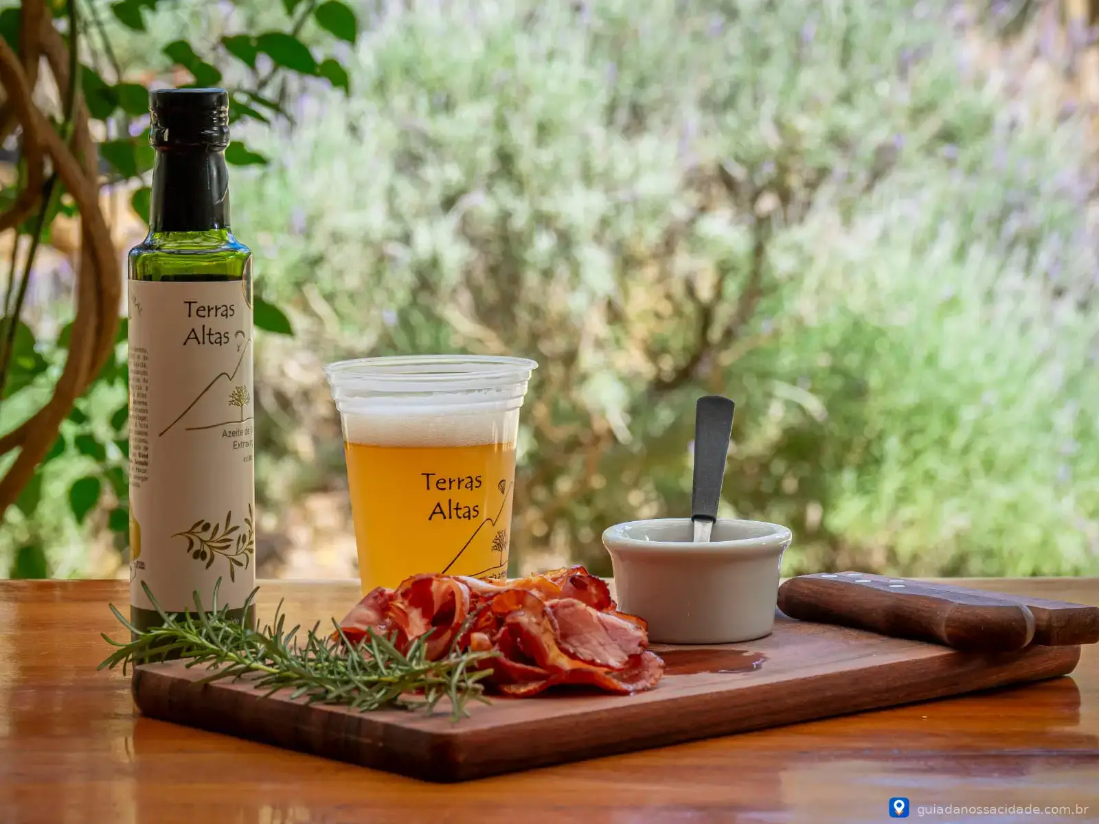 Mesa com azeite, copo de cerveja, tábuas de frios e ervas, ao ar livre com vegetação ao fundo.