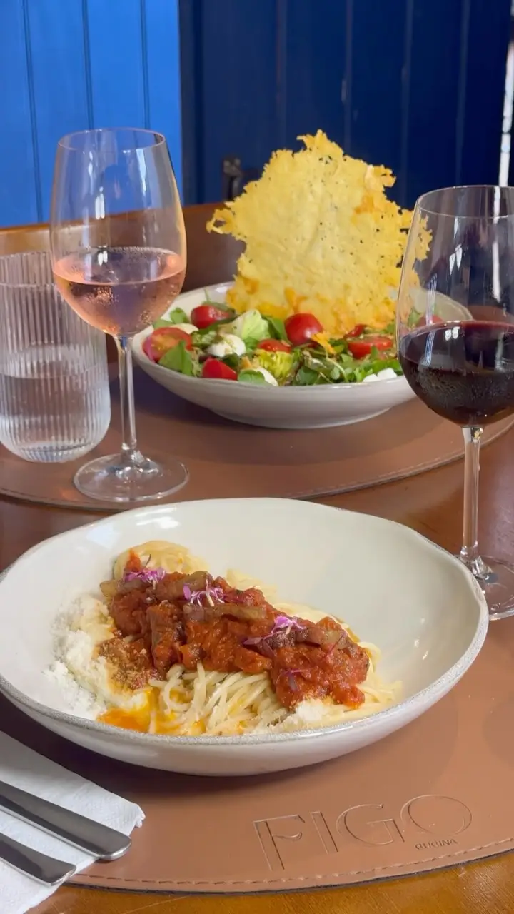 Prato de espaguete com molho, taças de vinho e salada ao fundo.