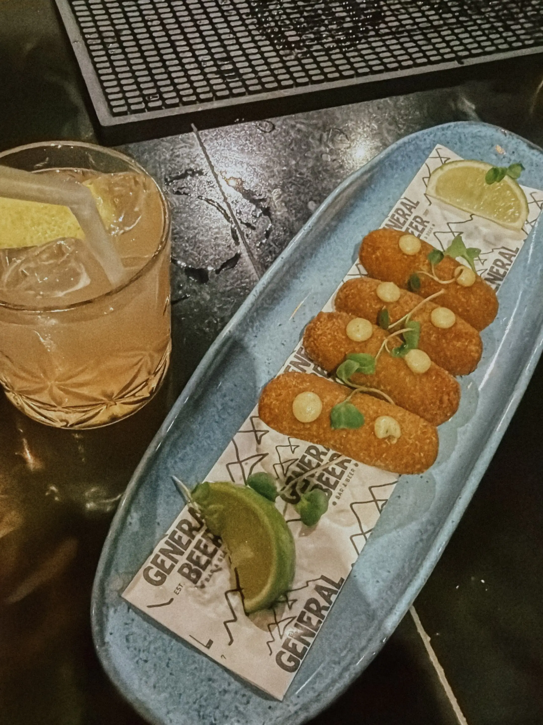 Bebida com gelo e fatia de limão ao lado de bolinhos fritos em prato azul, decorados com maionese e ervas.