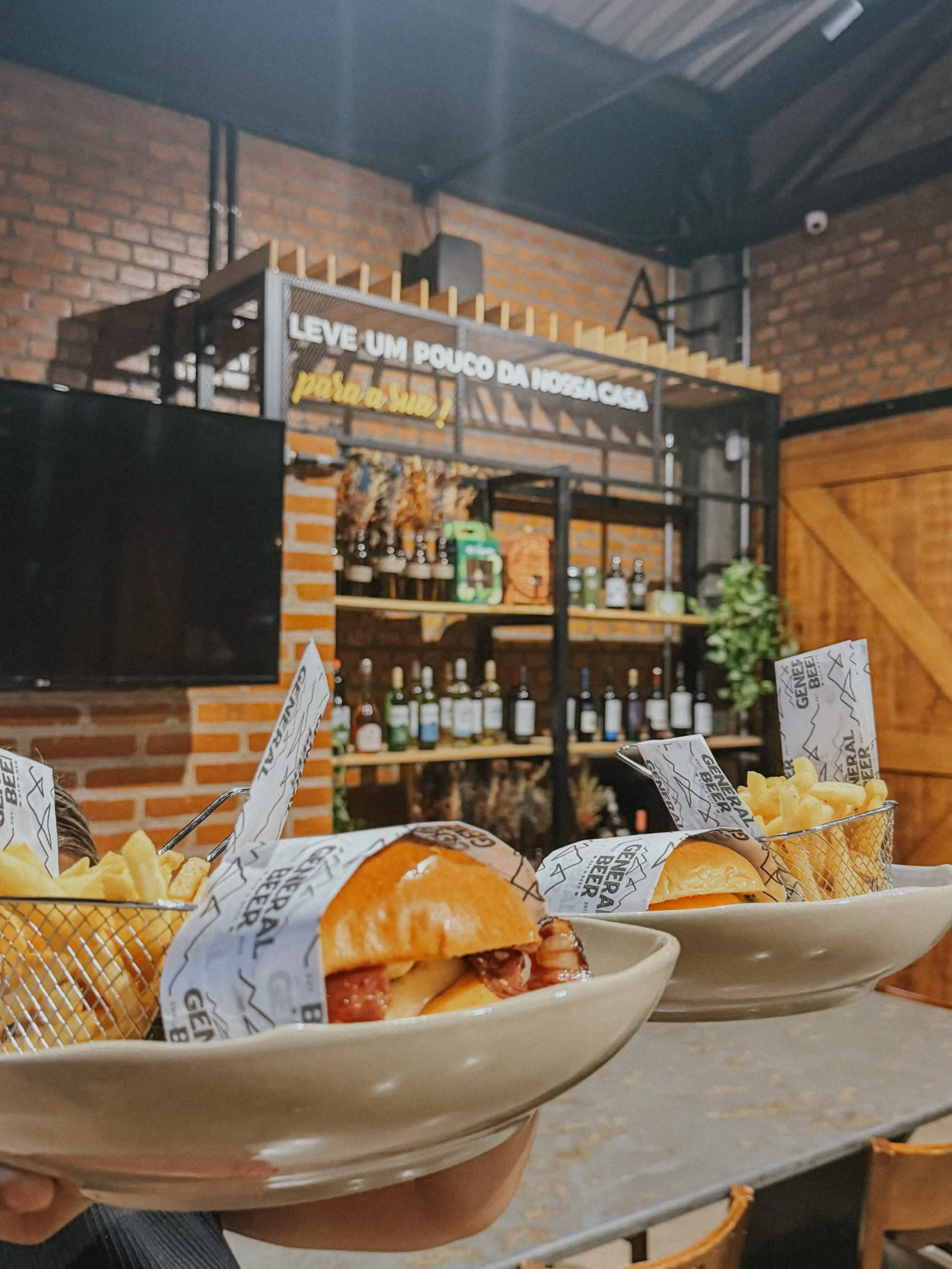 Pratos com hambúrgueres e batatas fritas em frente a estante com garrafas em restaurante rústico.