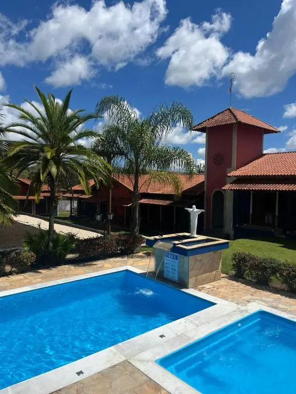 Área de lazer com piscina, estátua, palmeiras e construção com telhado vermelho ao fundo.