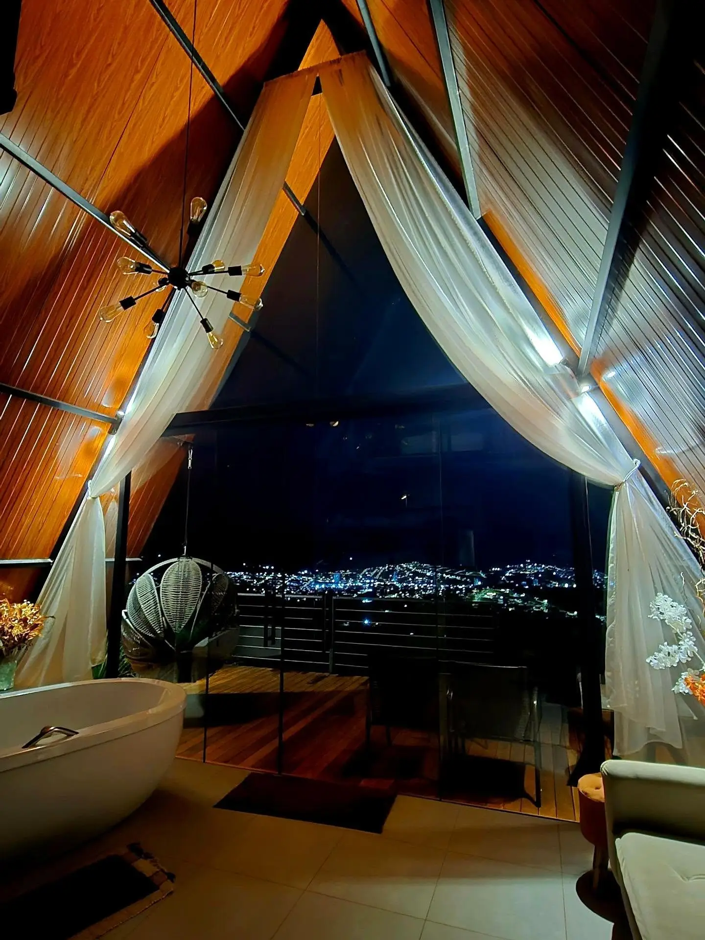 Sala com teto alto de madeira, cortinas brancas, banheira e vista noturna para cidade iluminada.