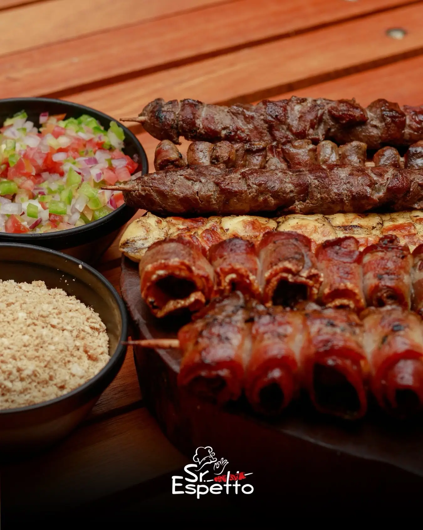 Espetinhos de carne e linguiça em tábua de madeira com vinagrete e farofa em bowls.