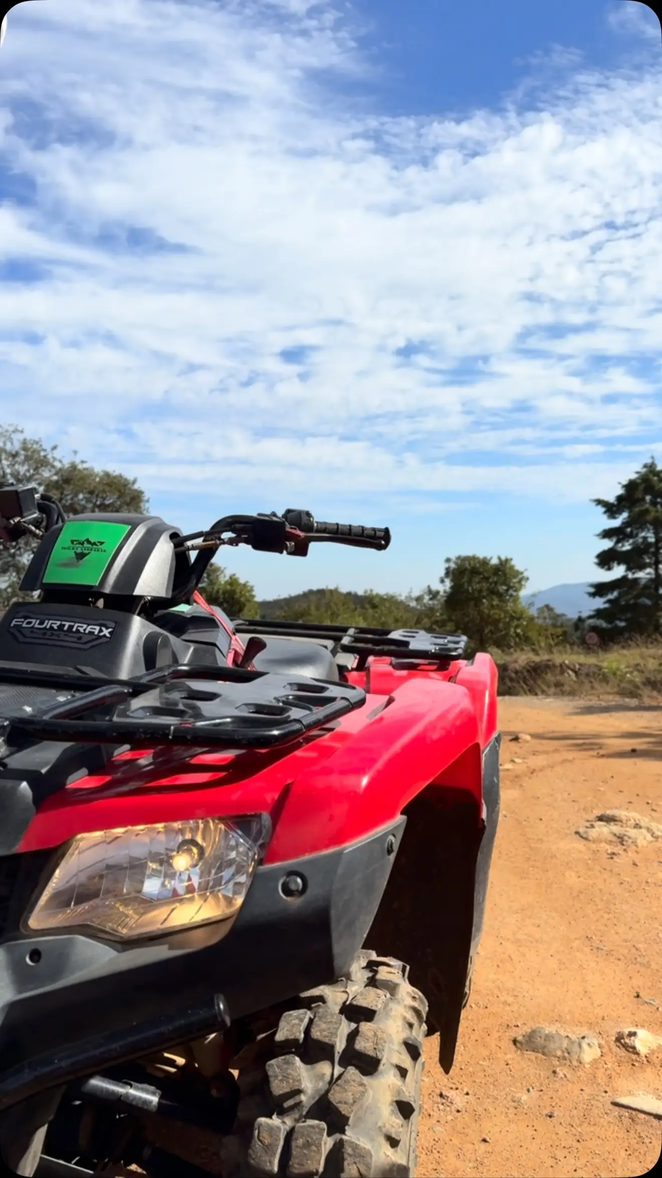 Quadriciclo vermelho em estrada de terra com céu azul e nuvens.
