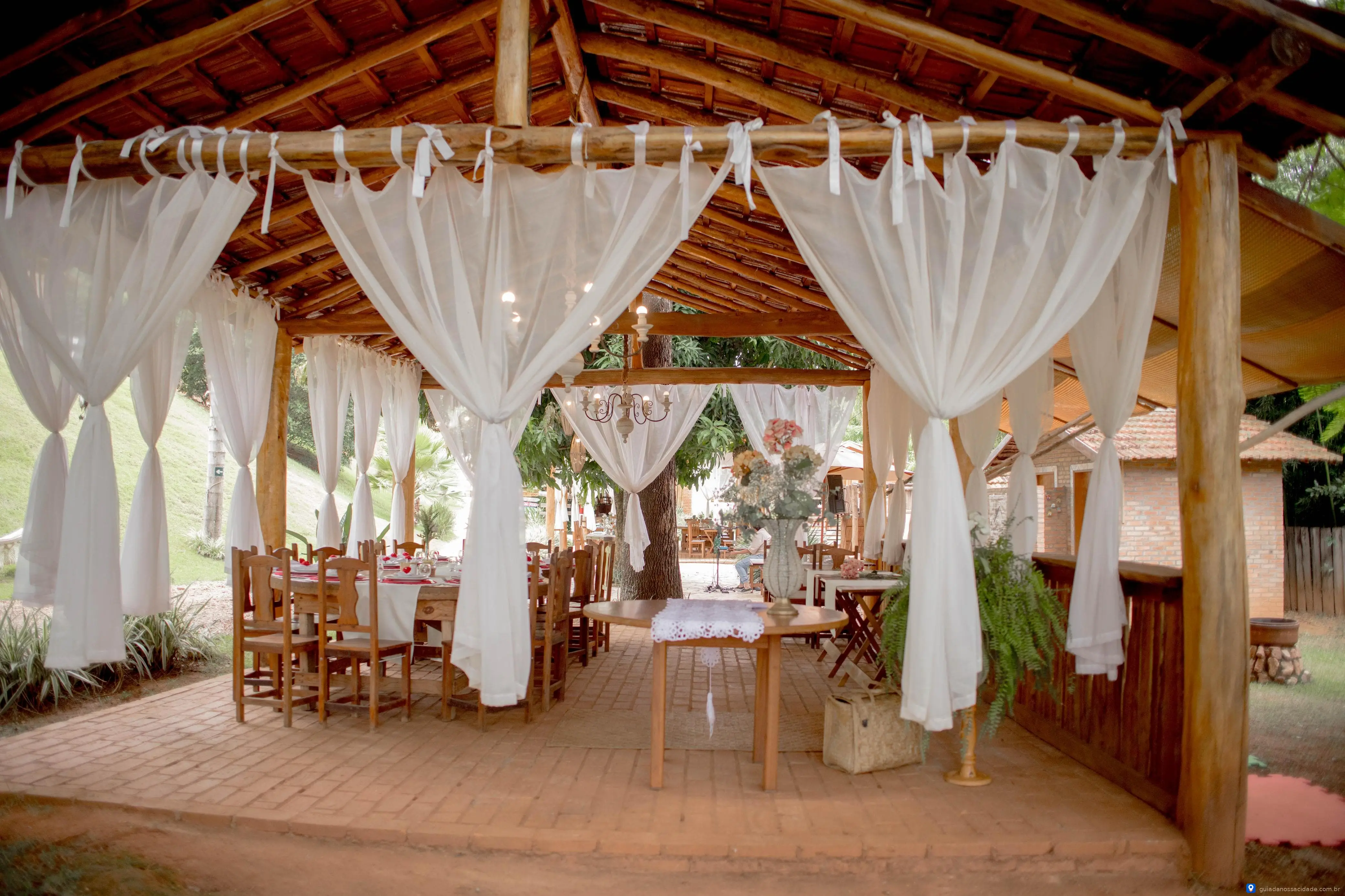 Varanda rústica de madeira com cortinas brancas, mesa, cadeiras e decoração floral.
