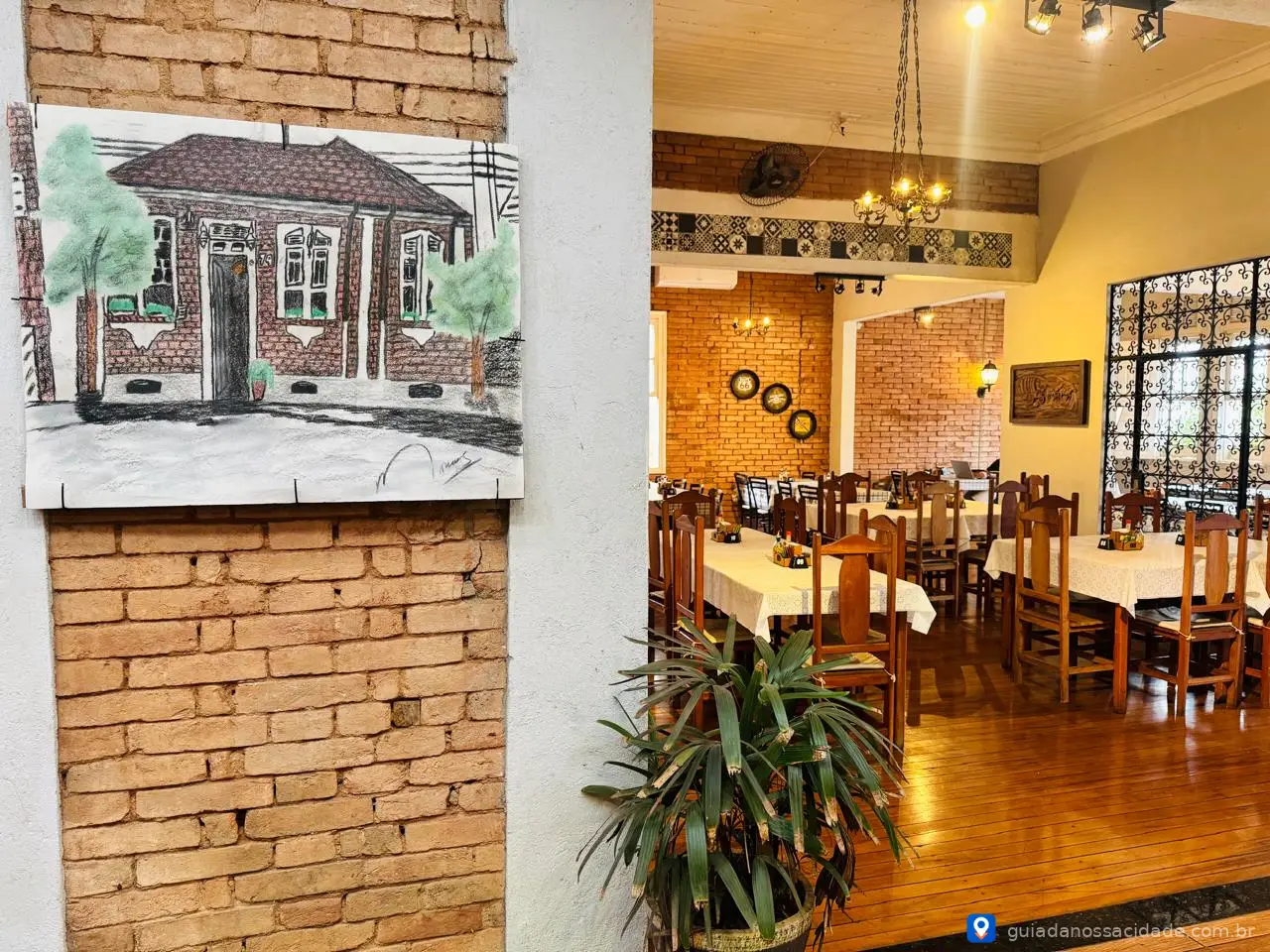 Sala de jantar rústica com mesas e cadeiras de madeira, piso de madeira e parede de tijolos, com quadro de casa antiga.