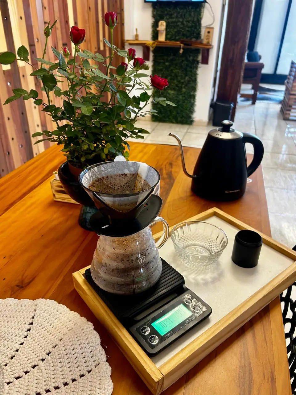 Mesa de madeira com cafeteira, bule preto, balança digital e vaso de rosas vermelhas.