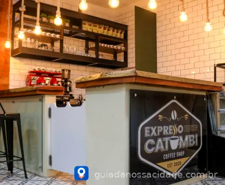Interior de cafeteria com balcão de madeira, letreiro Expresso Catumbi, máquina de café vermelha e luminárias pendentes.