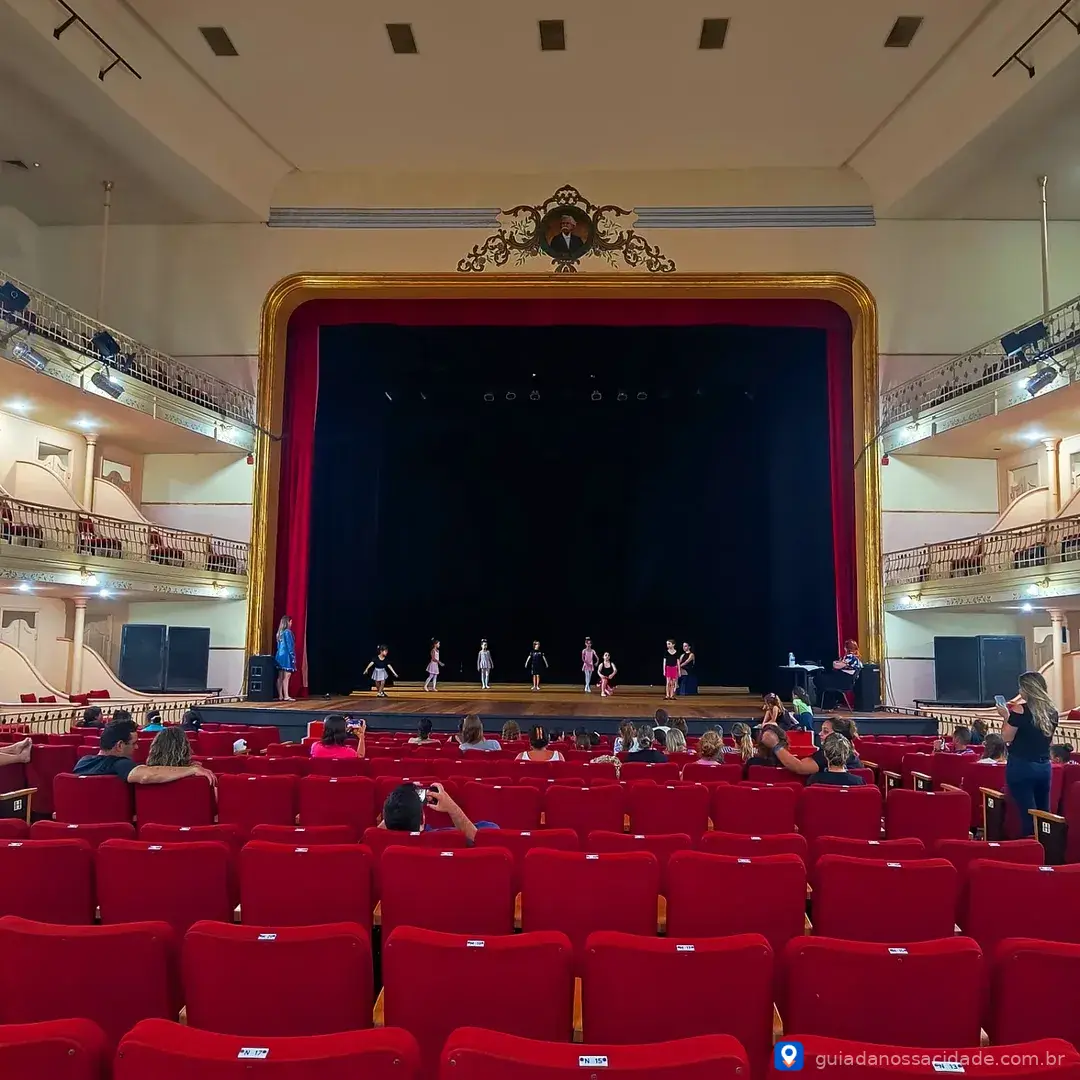 Crianças ensaiando dança no palco de um teatro com plateia de assentos vermelhos vazios.