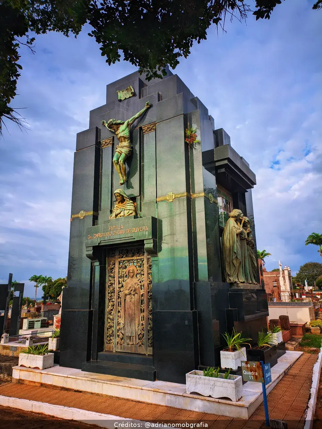 Mausoléu de mármore negro com esculturas religiosas e vegetação ao redor, sob céu parcialmente nublado.