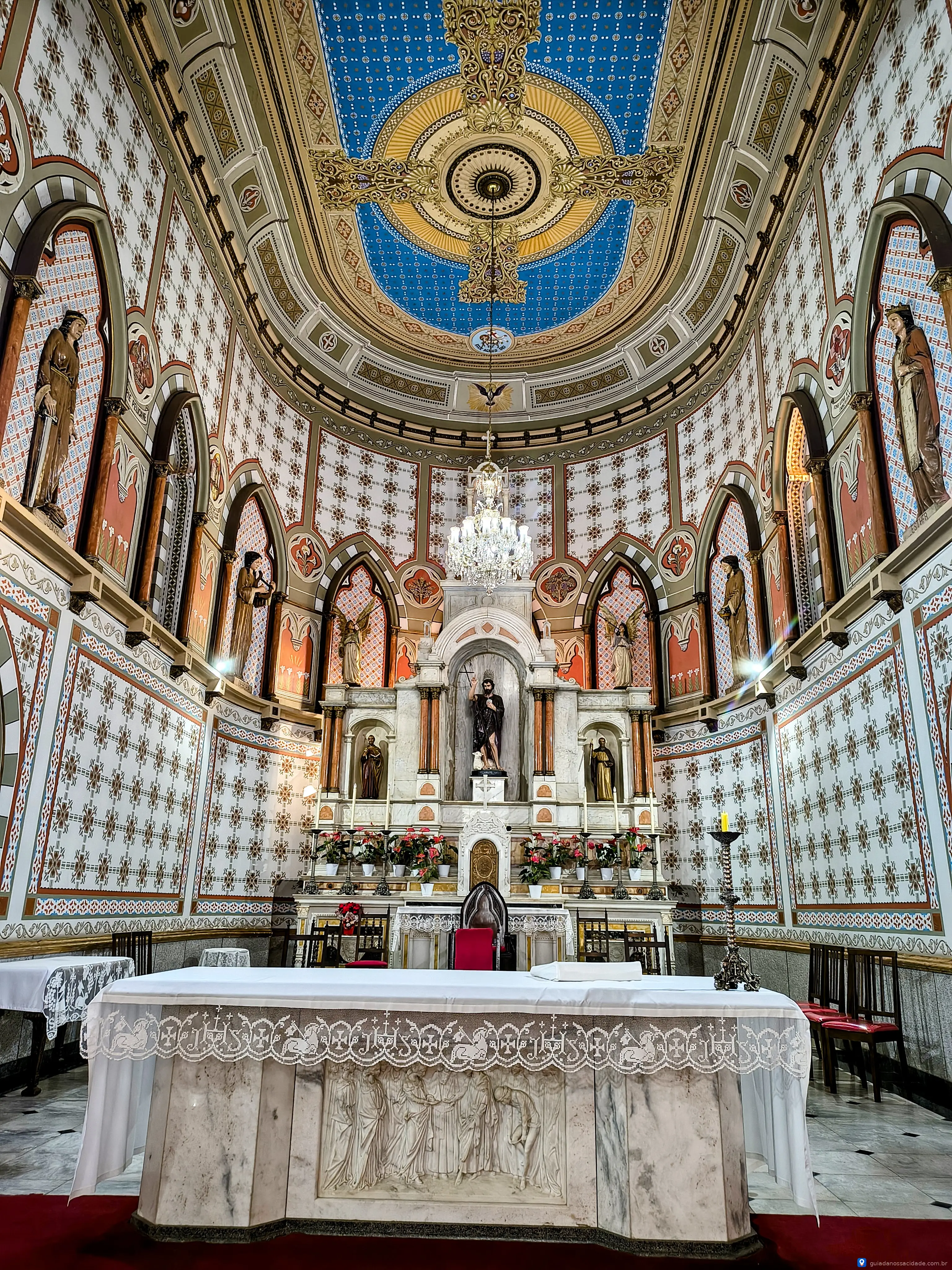 Interior de capela com altar ornado, teto azul decorado e estátuas religiosas em arco.