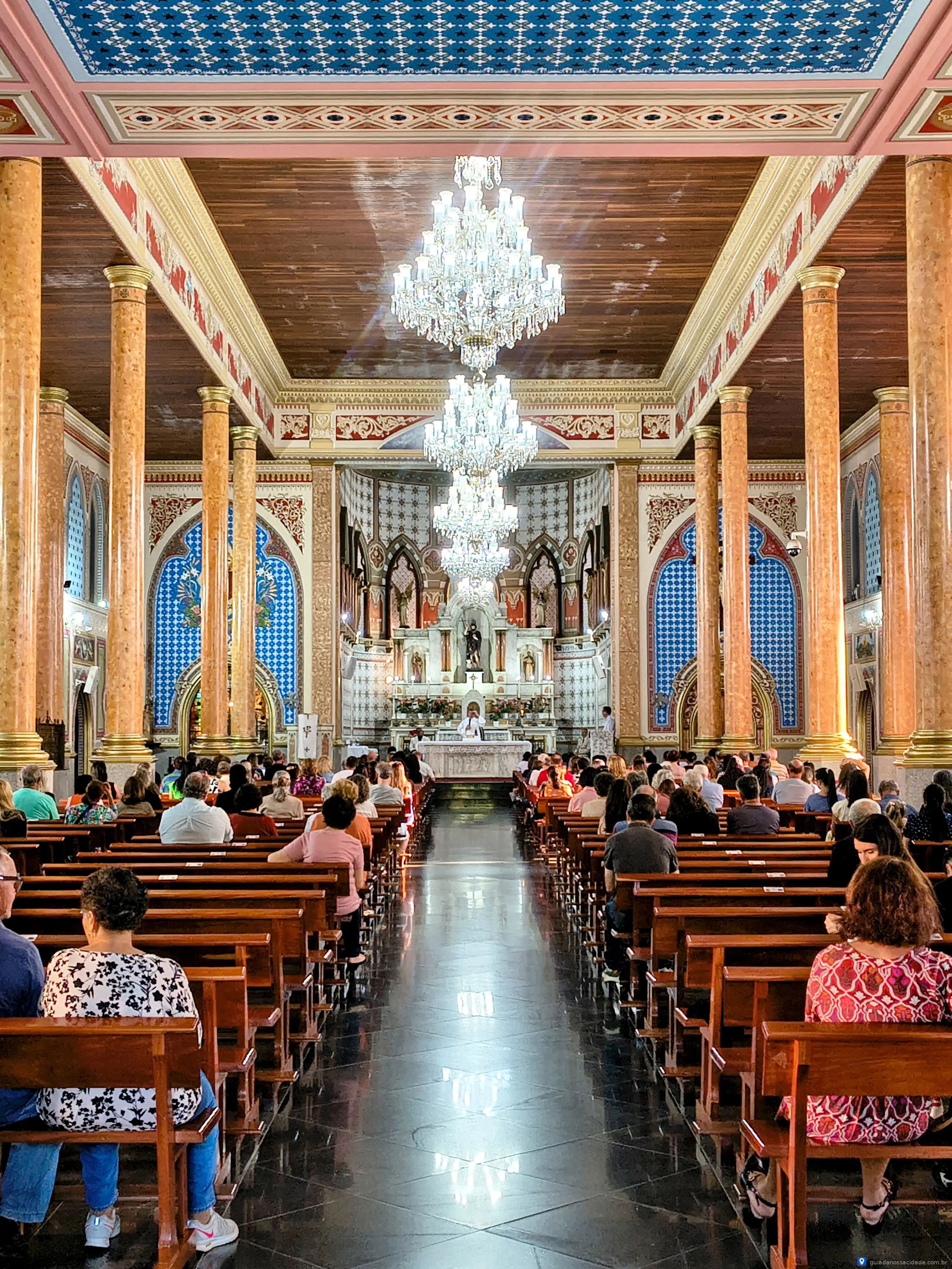 Interior de igreja com fileiras de bancos, vitrais decorativos e lustres, repleto de pessoas sentadas.
