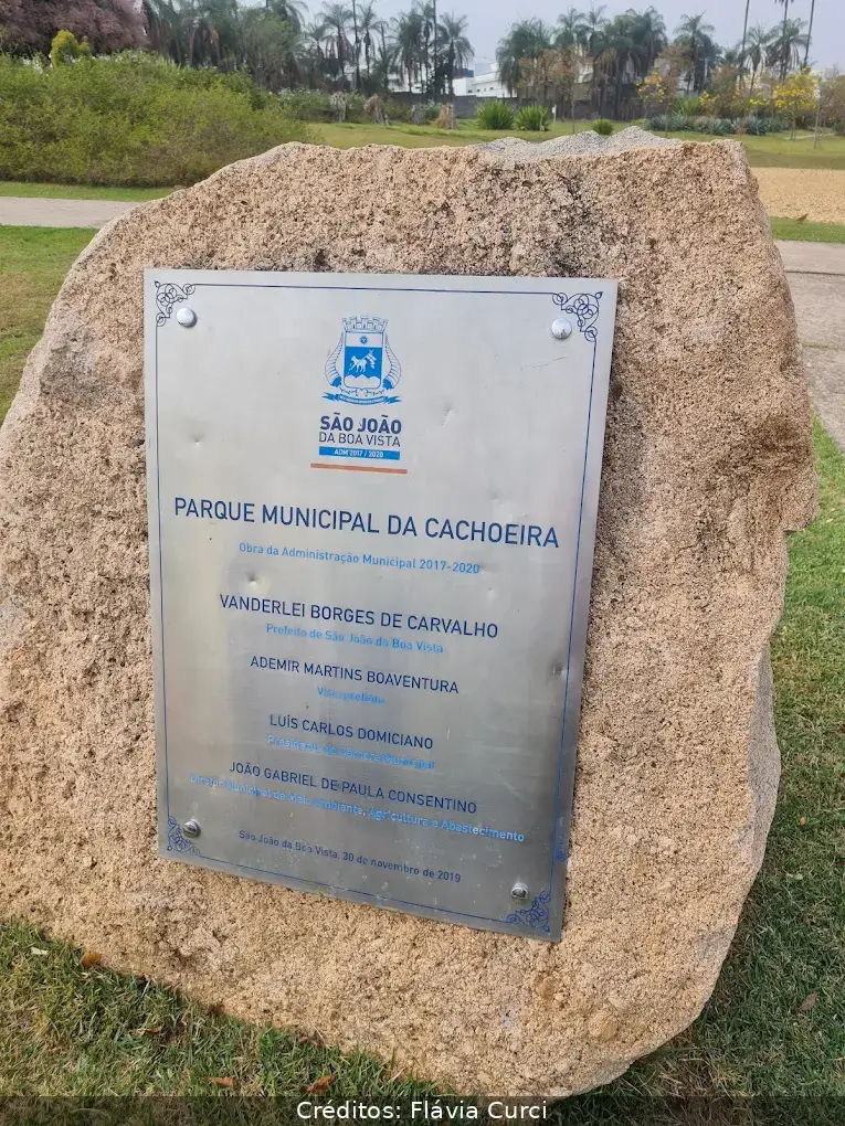 Placa de metal em pedra no Parque Municipal da Cachoeira em São João da Boa Vista, cercada por grama e árvores ao fundo.