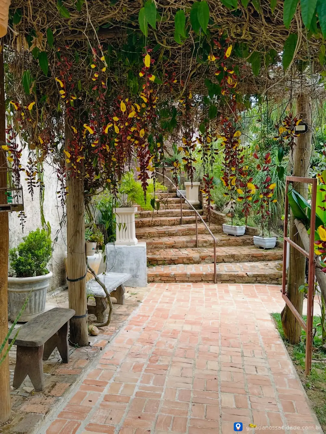 Corredor com piso de tijolos, plantas pendentes e escada de pedras ao fundo.